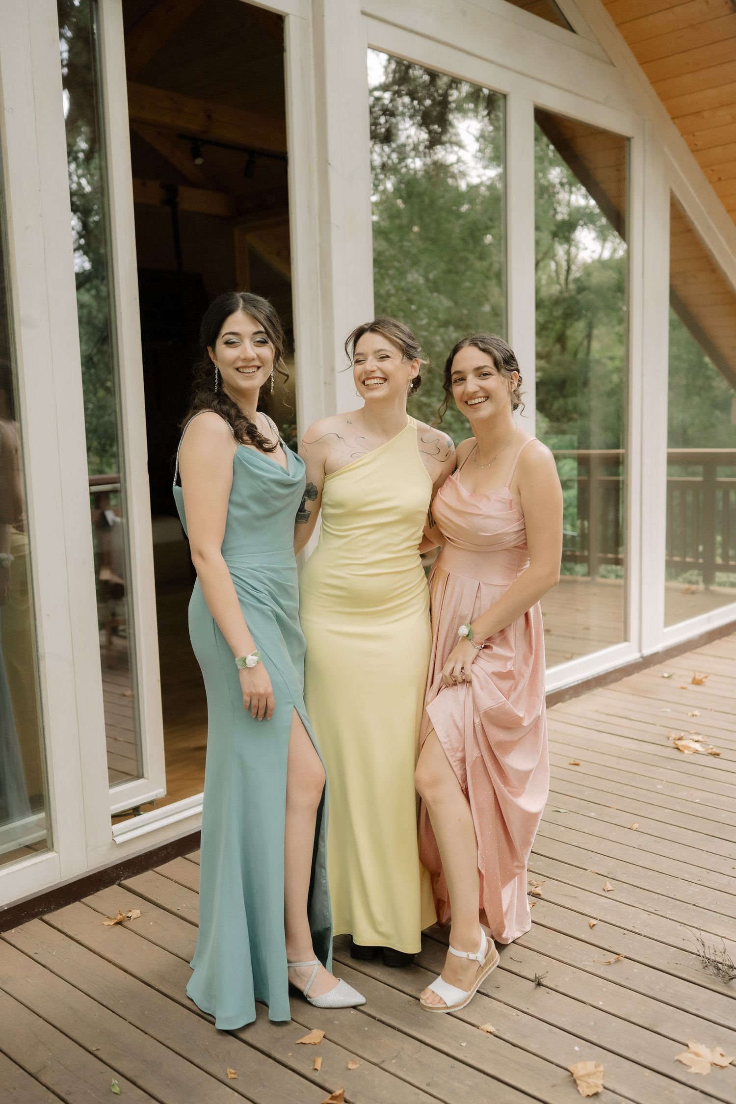 Three people in pastel dresses of mint, yellow and pink pose together on a wooden deck with large windows behind them.