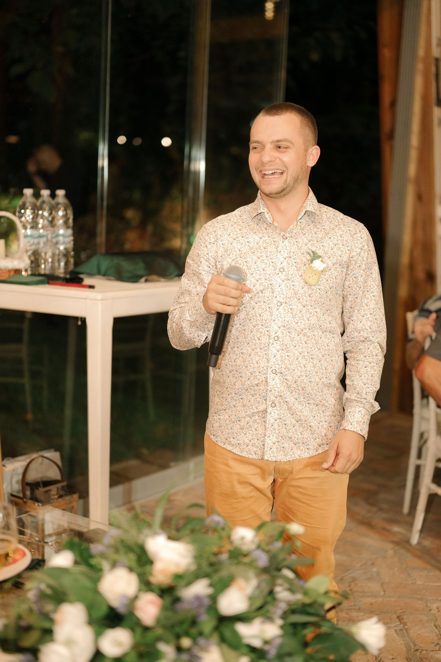 A person in a patterned button-down shirt and khaki pants stands smiling at an evening event with floral decorations.