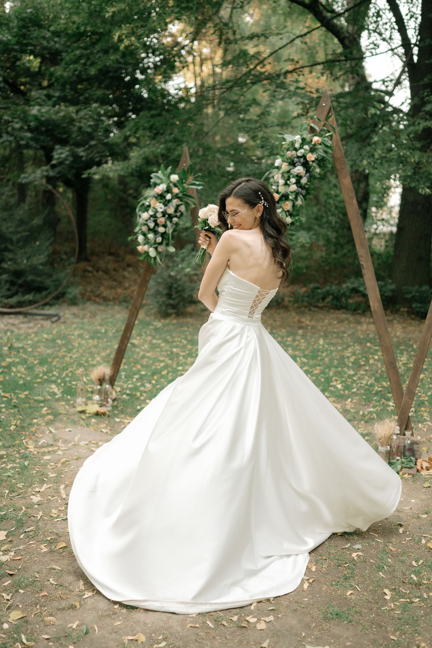 A bride in a flowing white satin ballgown twirls gracefully in a wooded outdoor setting during a wedding photoshoot.