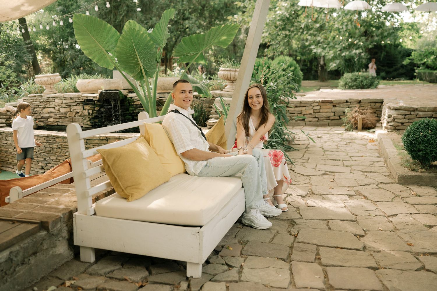 Two people relax together on a beige outdoor chaise lounge on a stone patio surrounded by lush greenery.