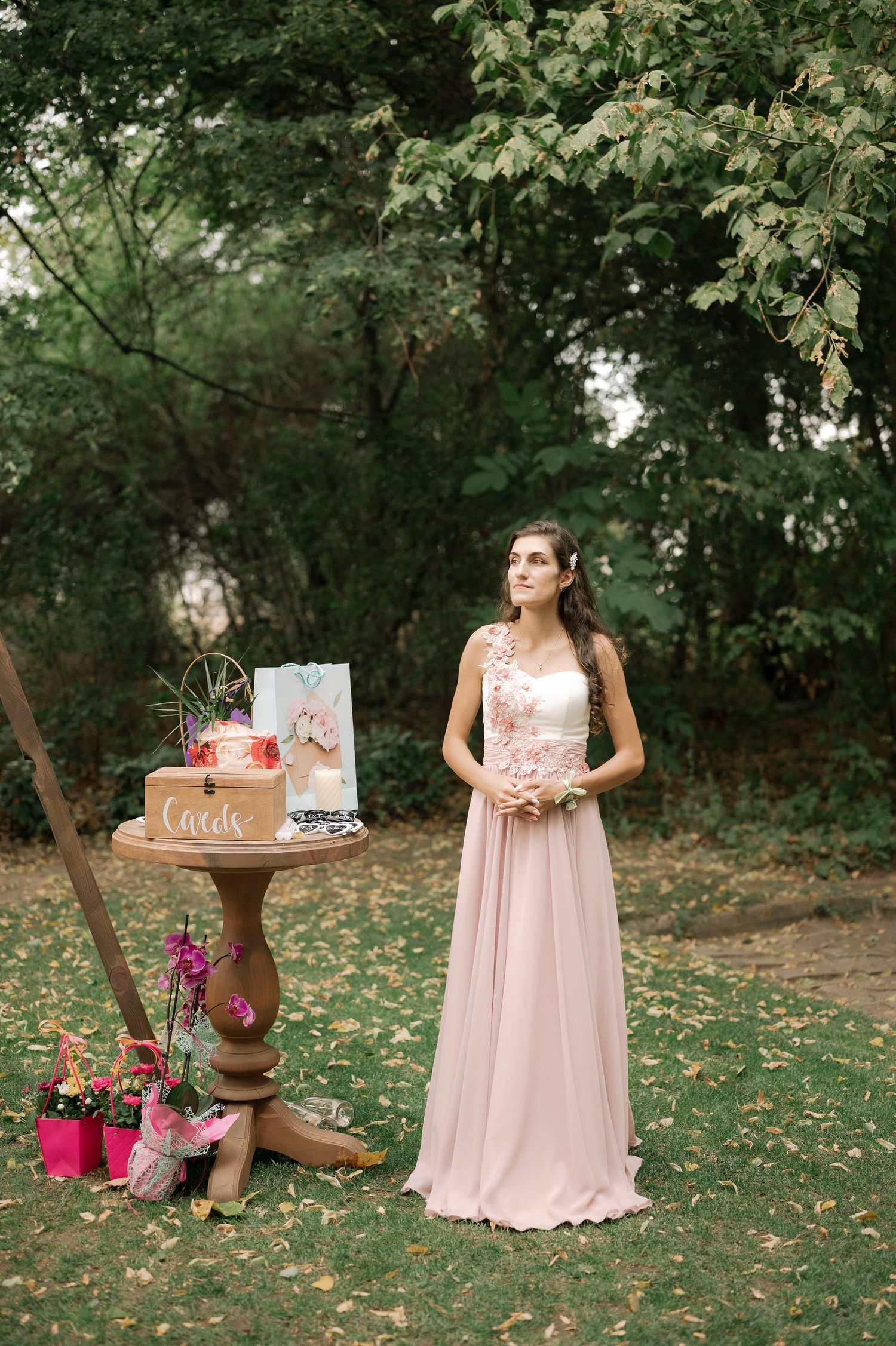 A pink floral wedding setup in a woodland garden with a rustic wooden table displaying cake and decorations.