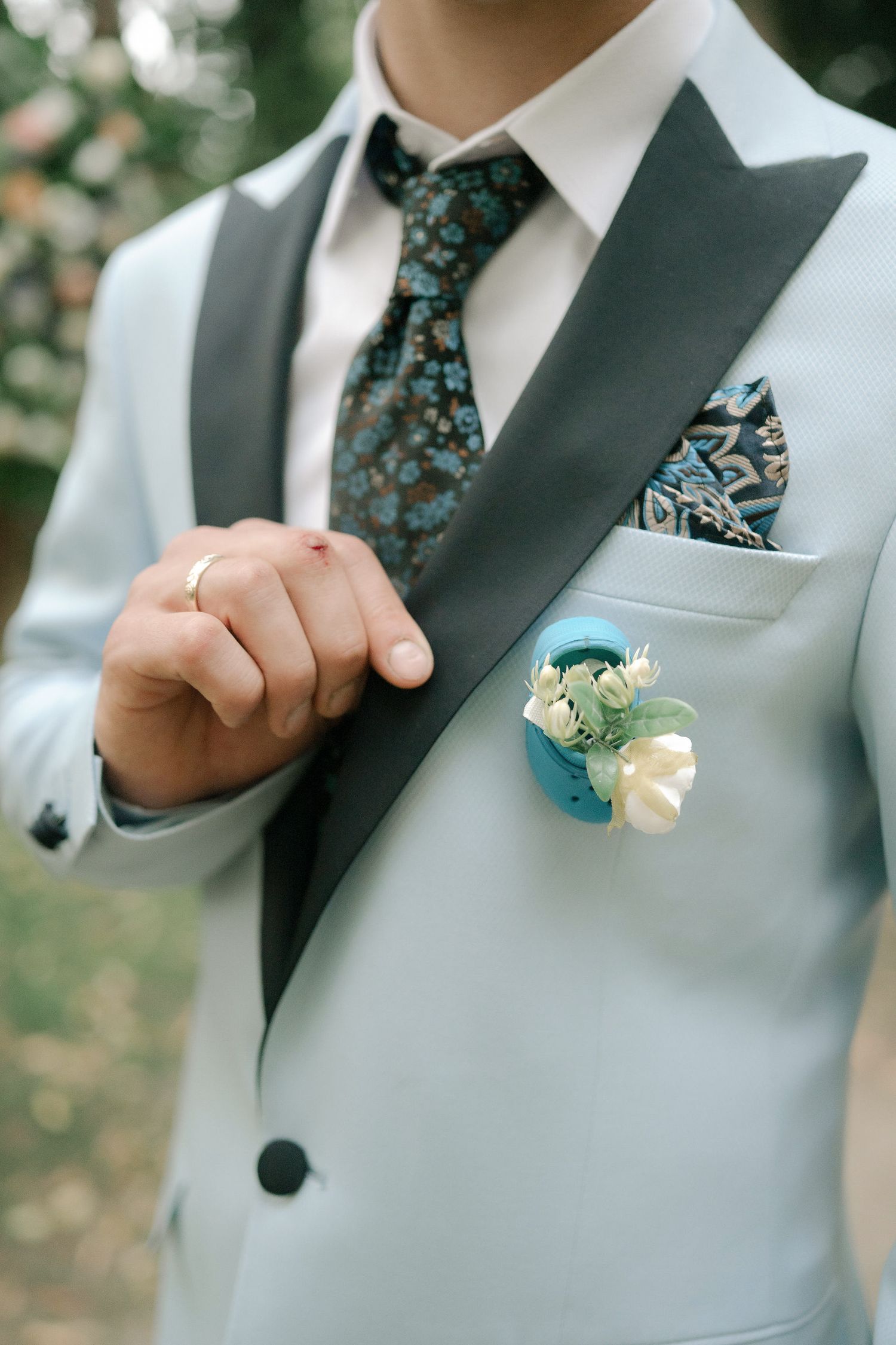 Close-up details of a light gray suit jacket with teal and black patterned tie and floral boutonniere.