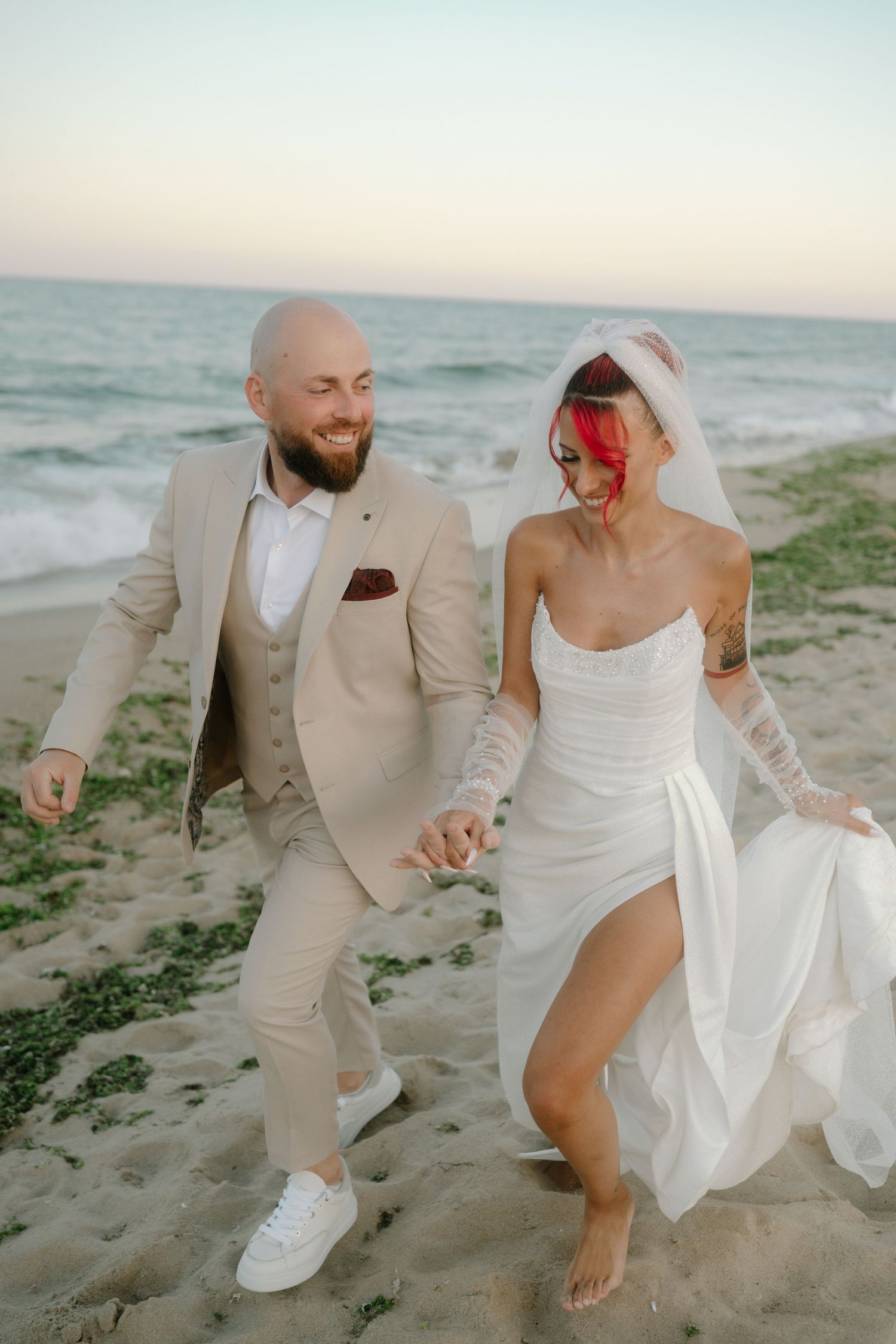 Newlywed couple playfully dancing and running together on a sandy beach at sunset in beige and white attire.