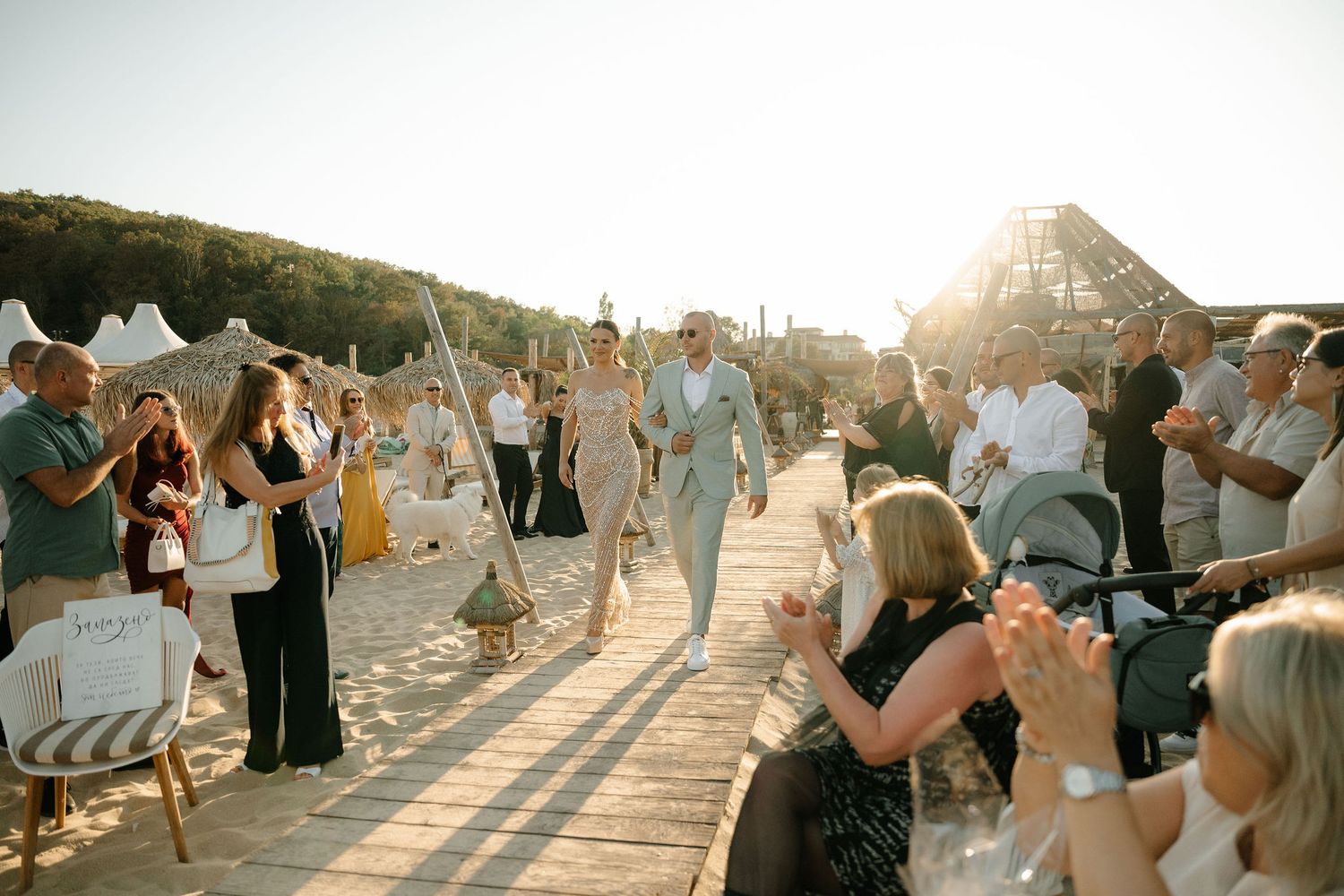 Wedding guests gather on a sandy beach at sunset as a couple walks down the aisle during an outdoor ceremony.