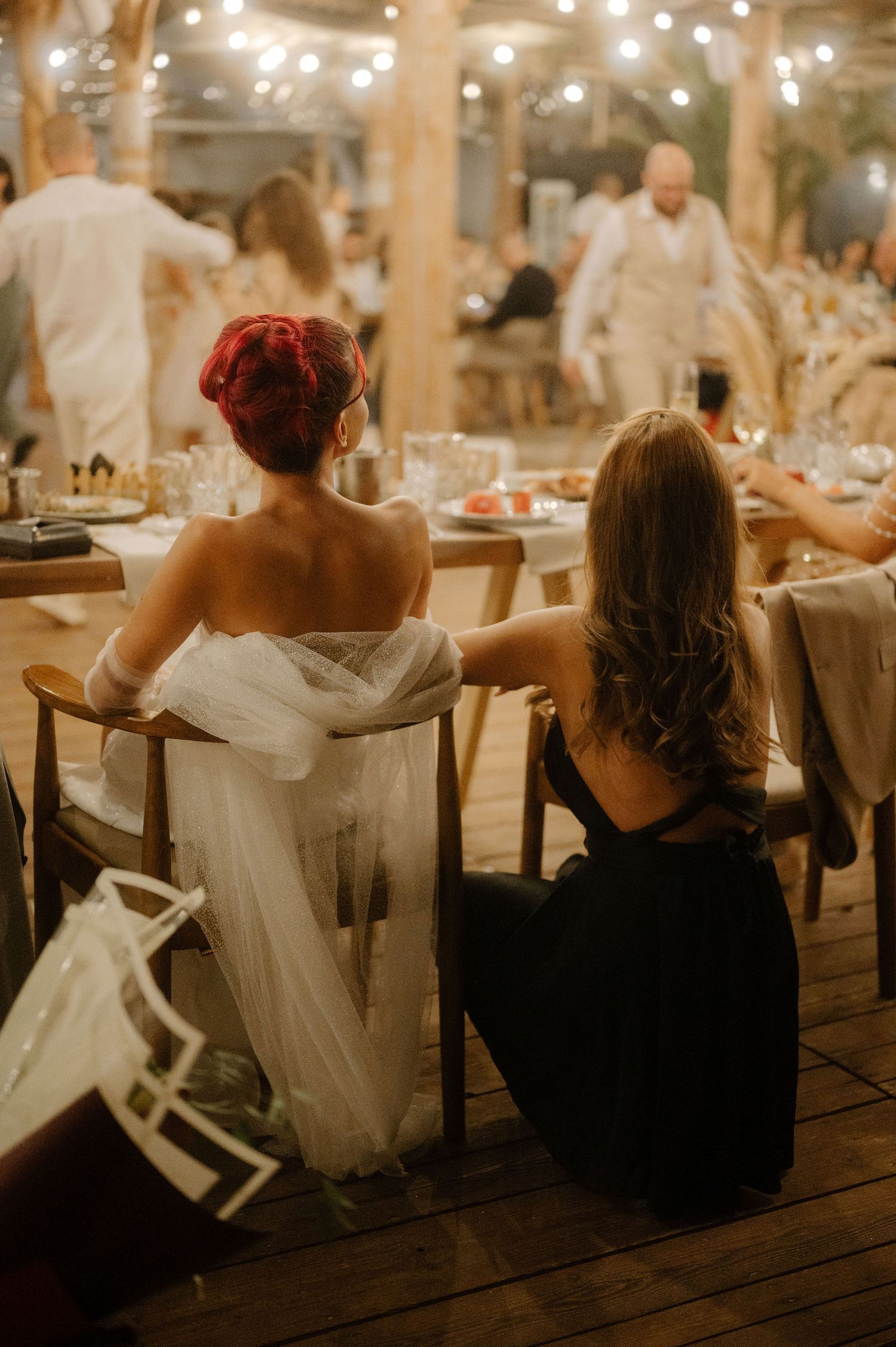 Two guests seated at an elegant wedding reception with string lights overhead and tables decorated for the celebration.