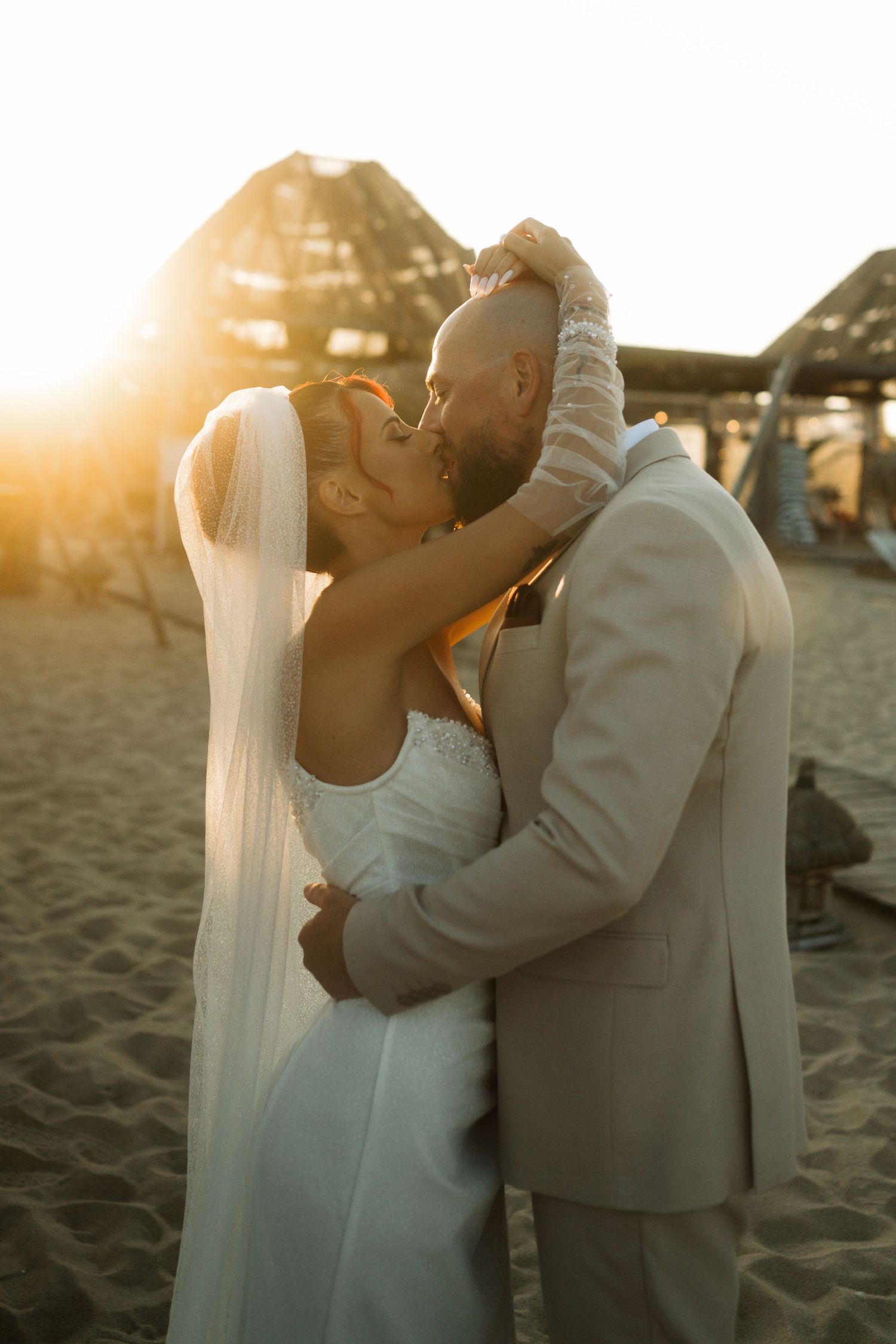 Newlywed couple shares romantic kiss on beach at sunset with golden light glowing behind them.
