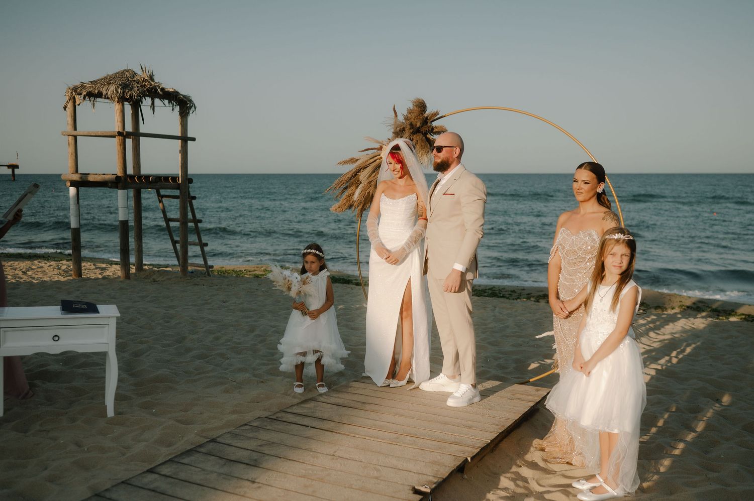 People in white outfits gather on a wooden pier by the ocean during a beach wedding ceremony at sunset.