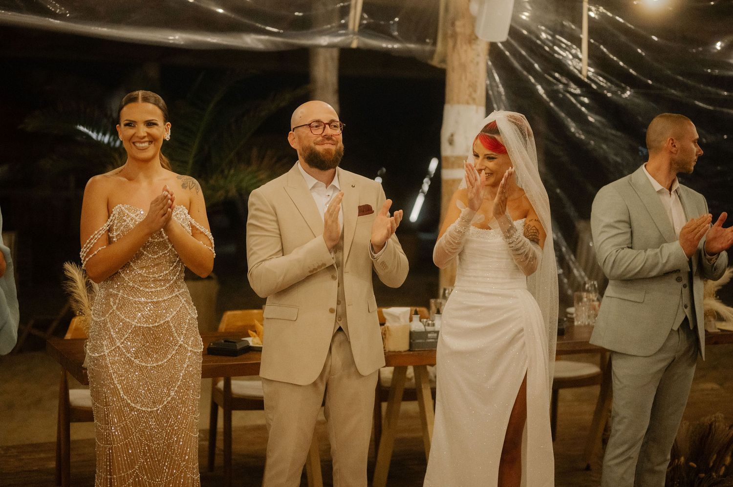 Wedding party members in formal attire applaud during an evening celebration at an outdoor venue.