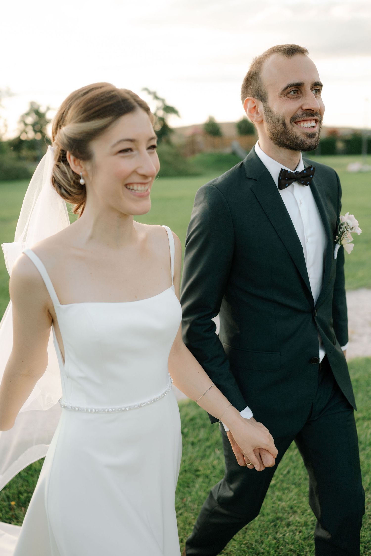 A happy bride and groom walk hand in hand through a grassy field on their wedding day.
