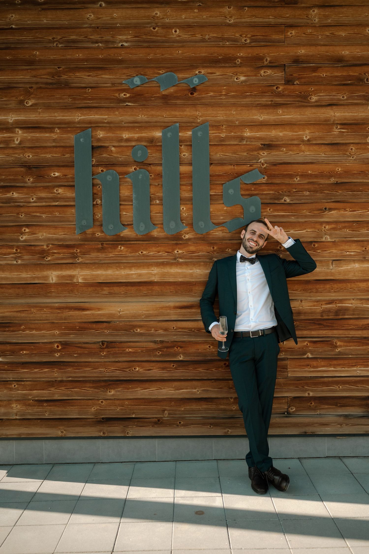 A person in a dark suit poses playfully against a wooden wall with text art installation.