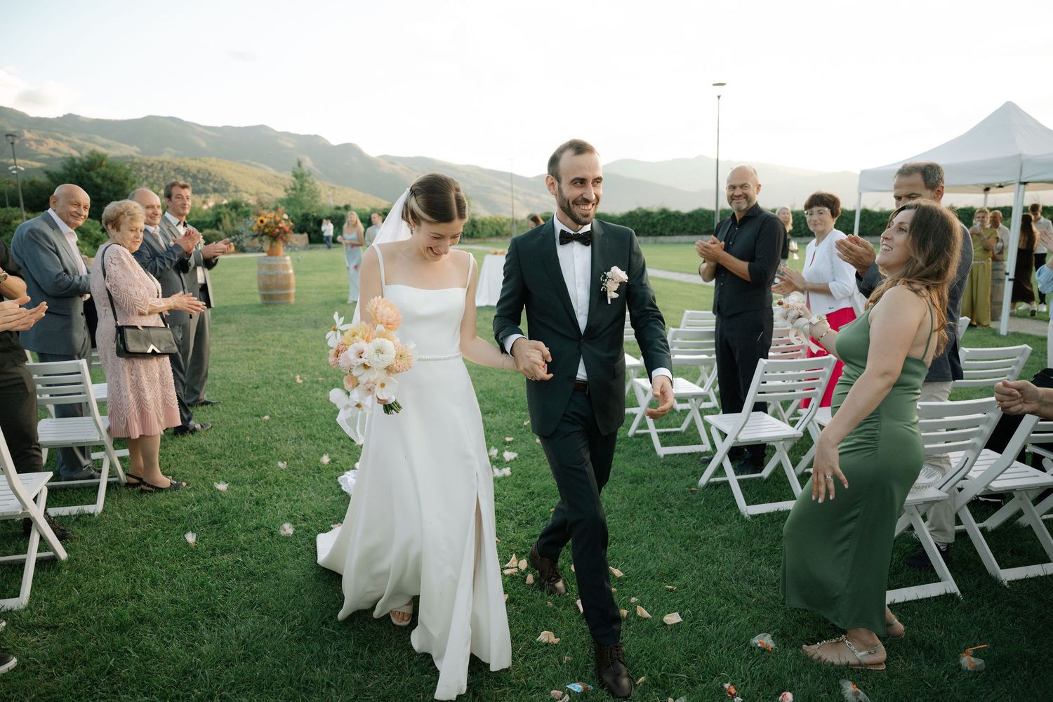 Wedding guests enjoy an outdoor reception with mountain views and white folding chairs on a lush green lawn.