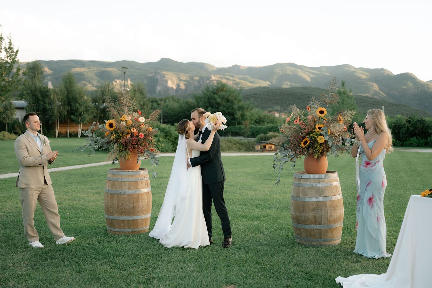 Wedding party gathers for outdoor celebration with wine barrels and mountain backdrop in rustic setting.