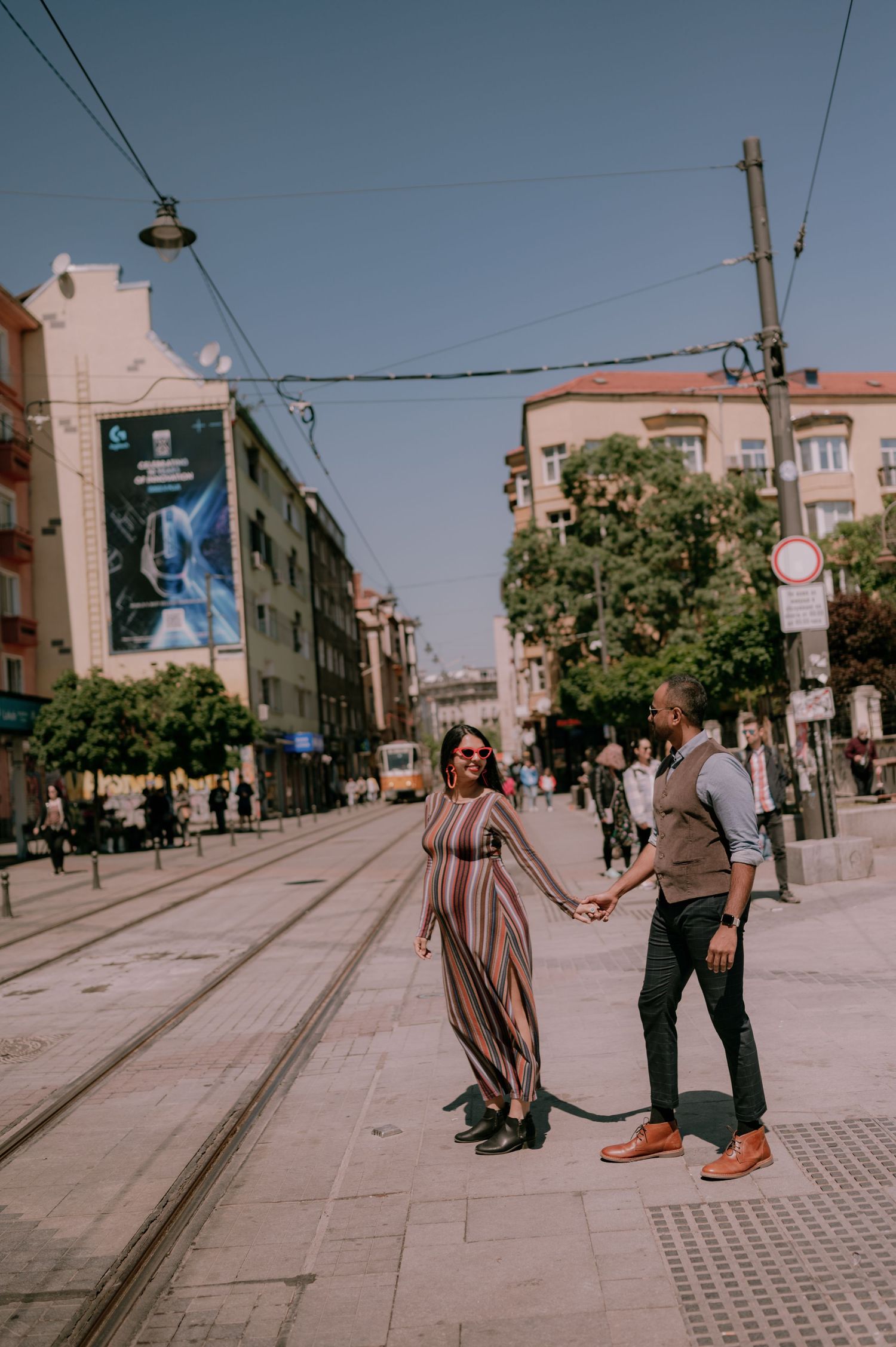 A couple holds hands on a sunny city street with tram tracks, the woman wearing a colorful striped maxi dress.