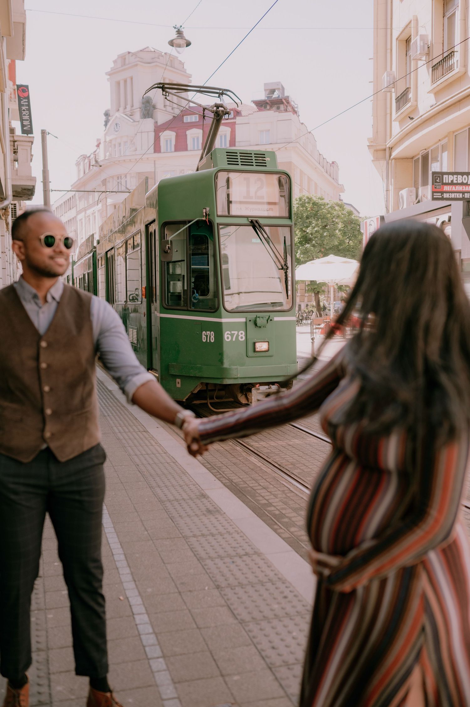 Couple holding hands on a city street with a green tram in the background on a sunny day.