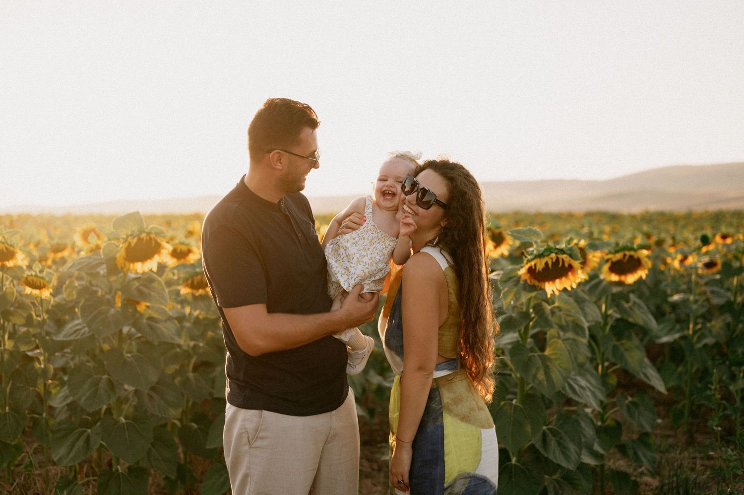 Family with baby posing in a sunflower field at golden hour sunset.