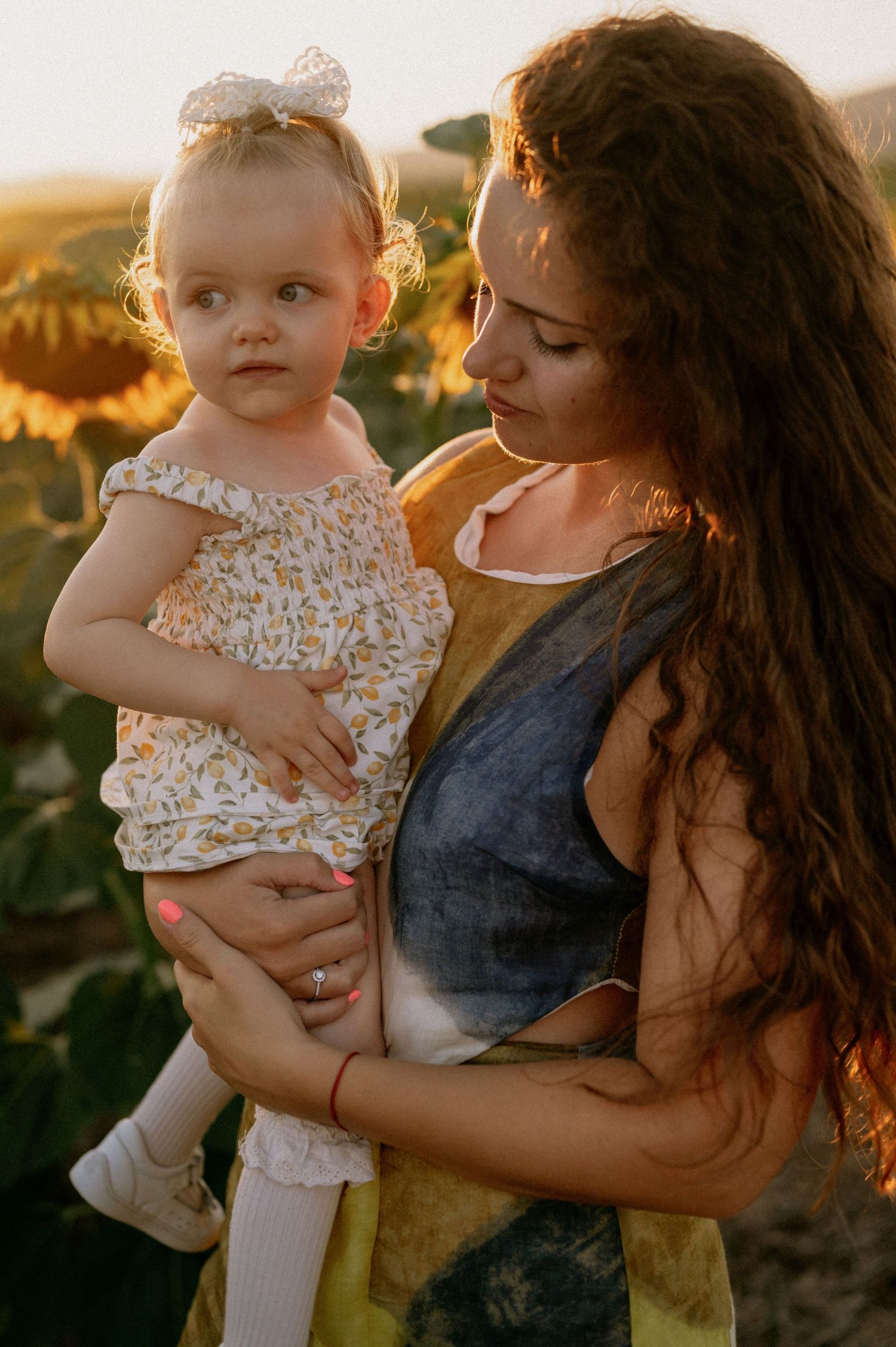 A mother holds her toddler daughter in a sunflower field at golden hour, both wearing floral outfits.