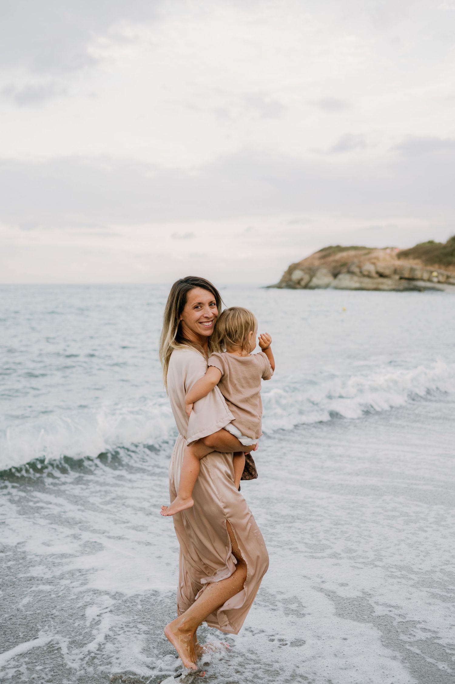 A smiling mother in a beige maxi dress holds her toddler while standing in ocean waves on a sandy beach.