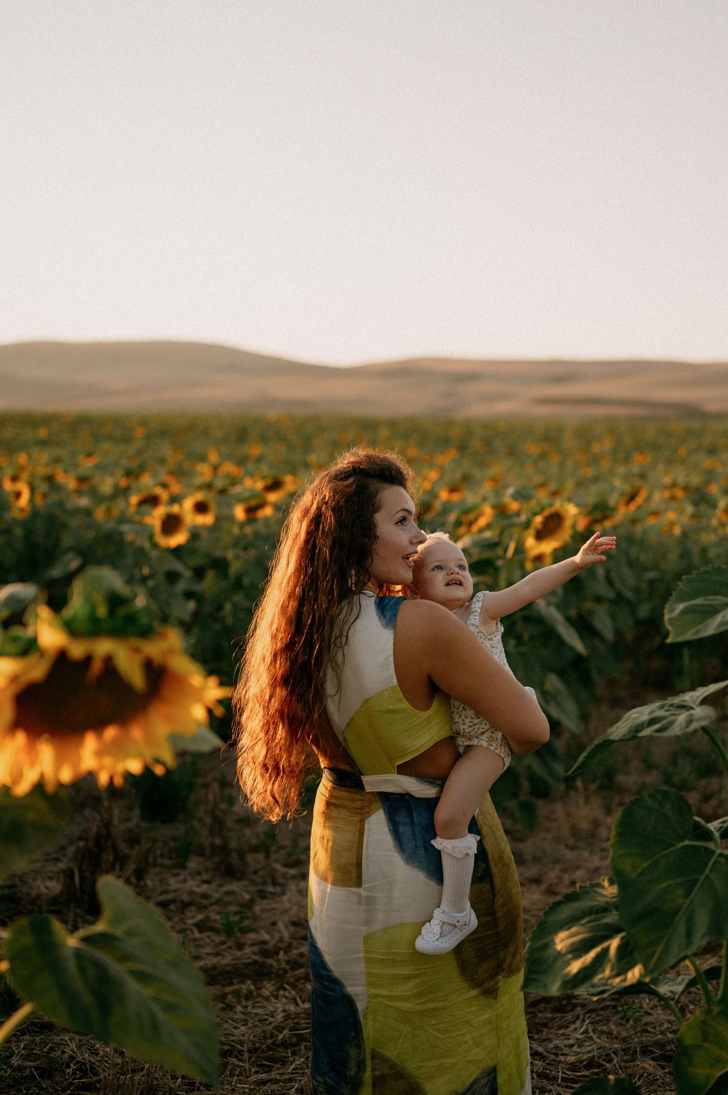 A mother holds her baby in a vast sunflower field at golden hour, as the child reaches out toward the horizon.