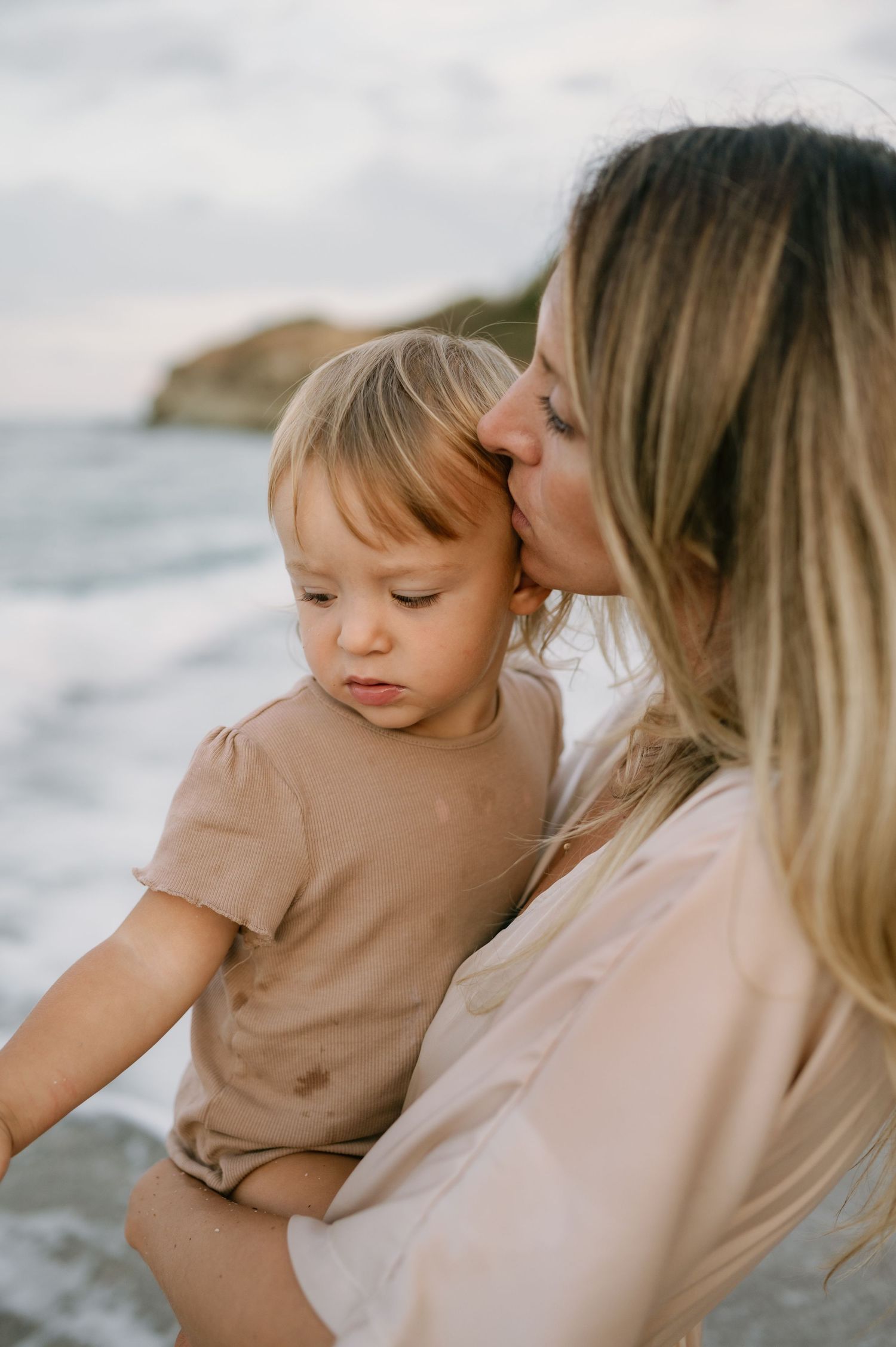 A mother tenderly kisses her toddler on the forehead while holding them at a scenic beach.