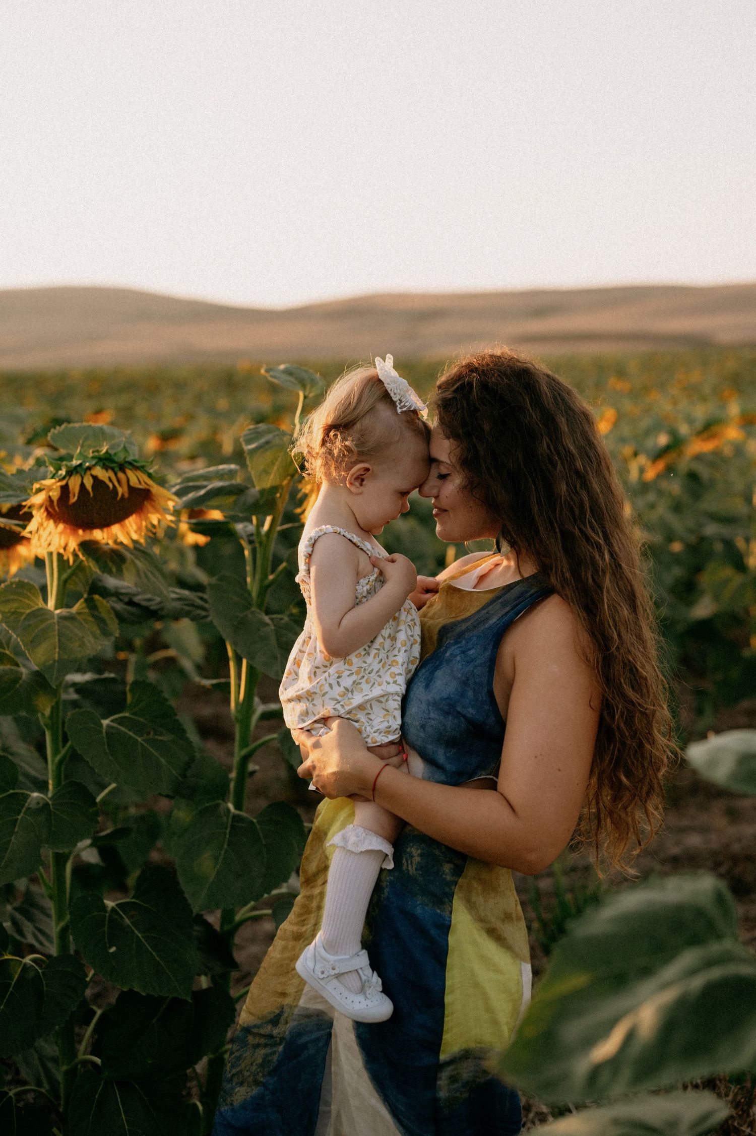 A mother holds her toddler daughter in a sunflower field at golden hour, their foreheads touching tenderly.