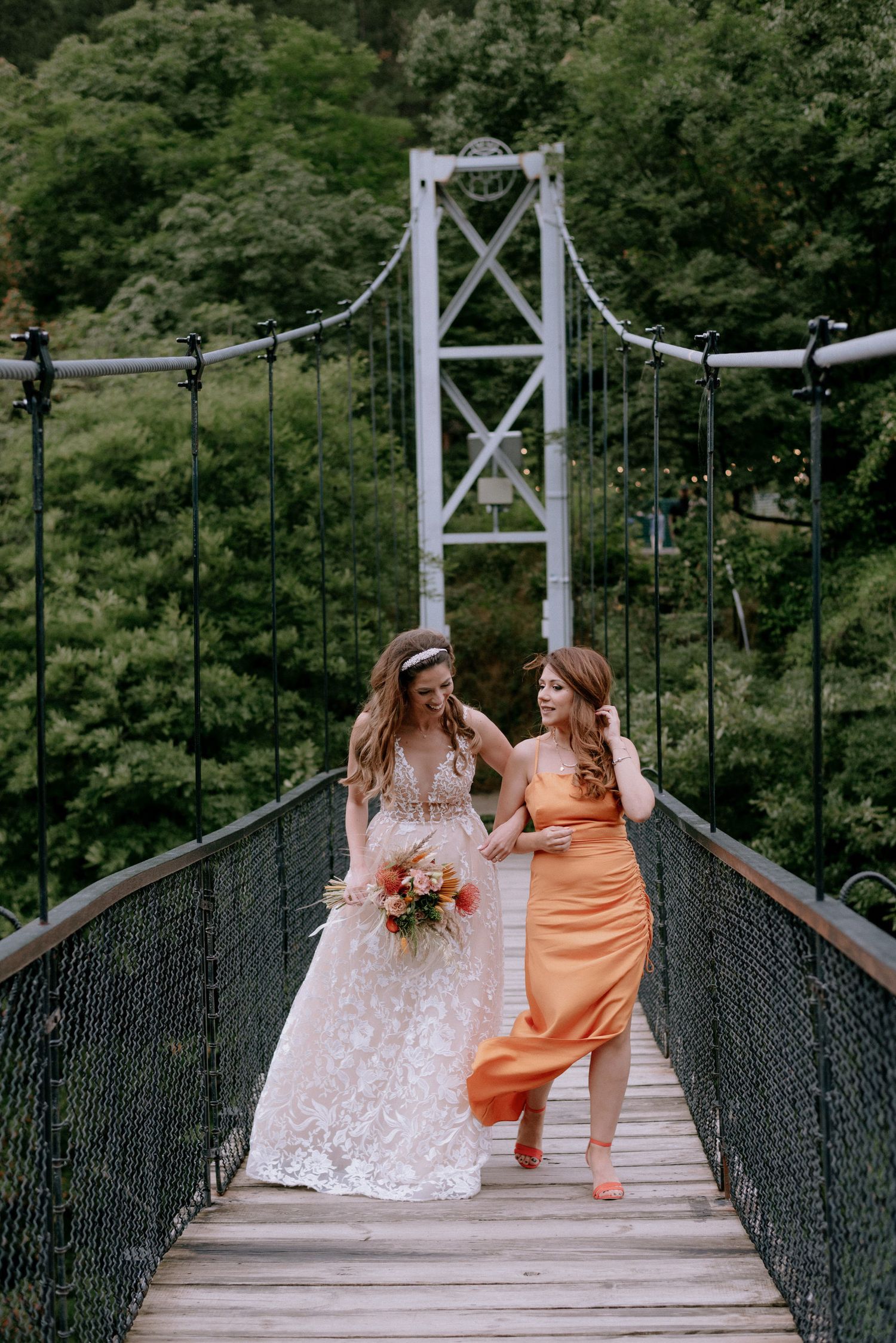Two people in colorful dresses share a joyful moment on a scenic suspension bridge surrounded by lush forest.
