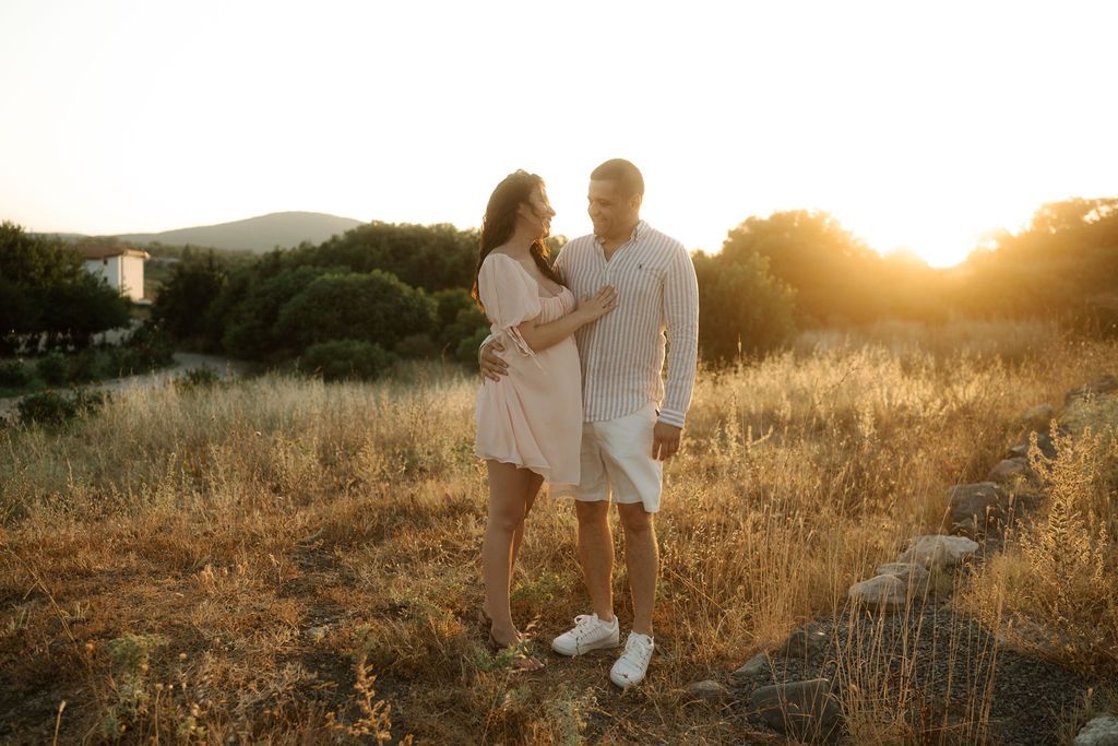 A couple stands together in a golden field at sunset wearing matching white summer outfits.