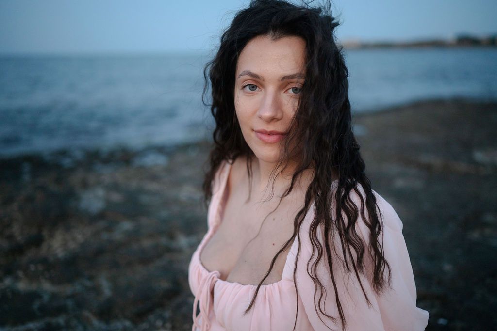 Portrait of someone with dark wavy hair wearing pink against a moody coastal background at dusk.