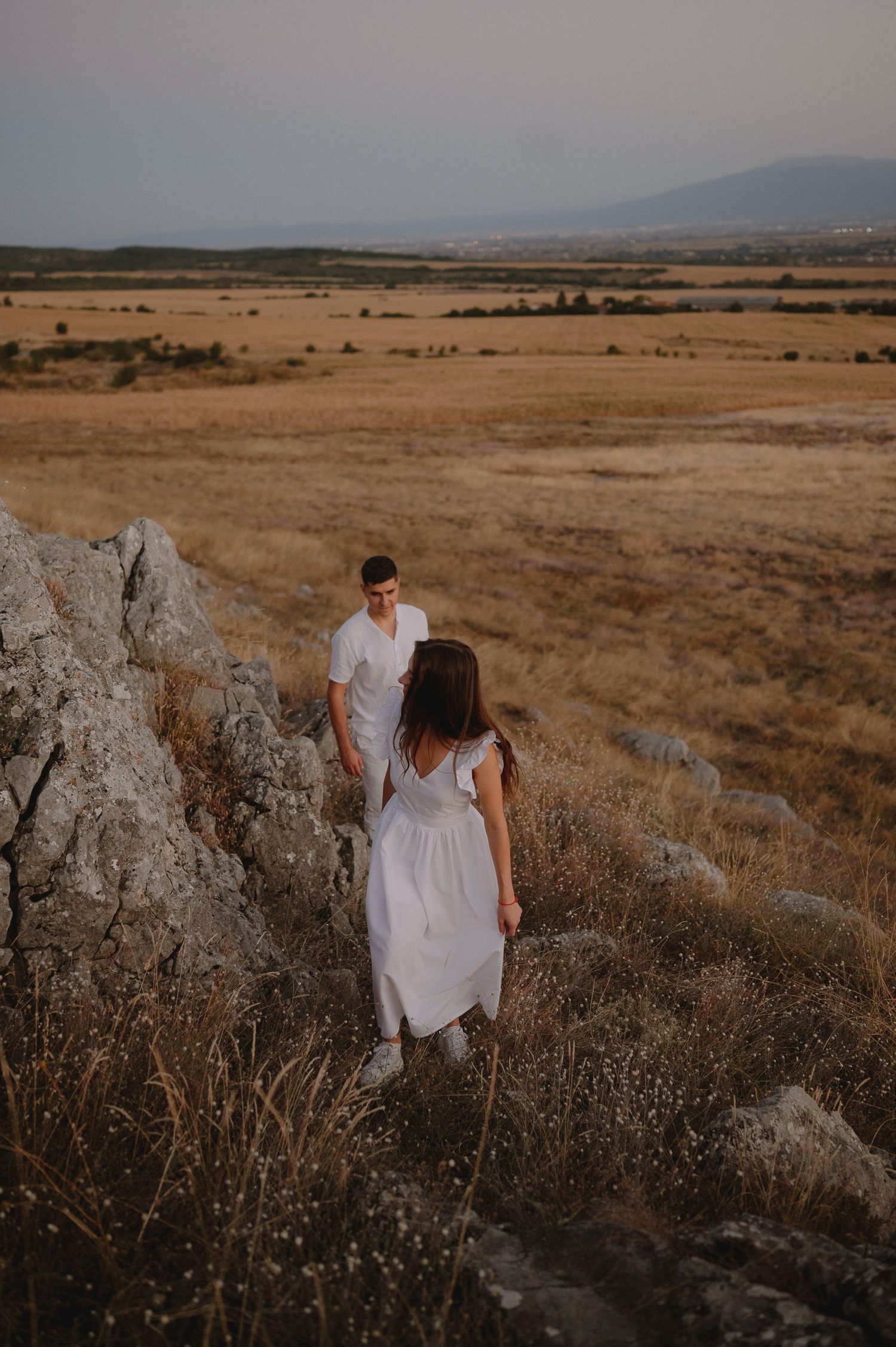 Couple walking through a golden field at sunset with mountains in the distance and large rocks in the foreground.