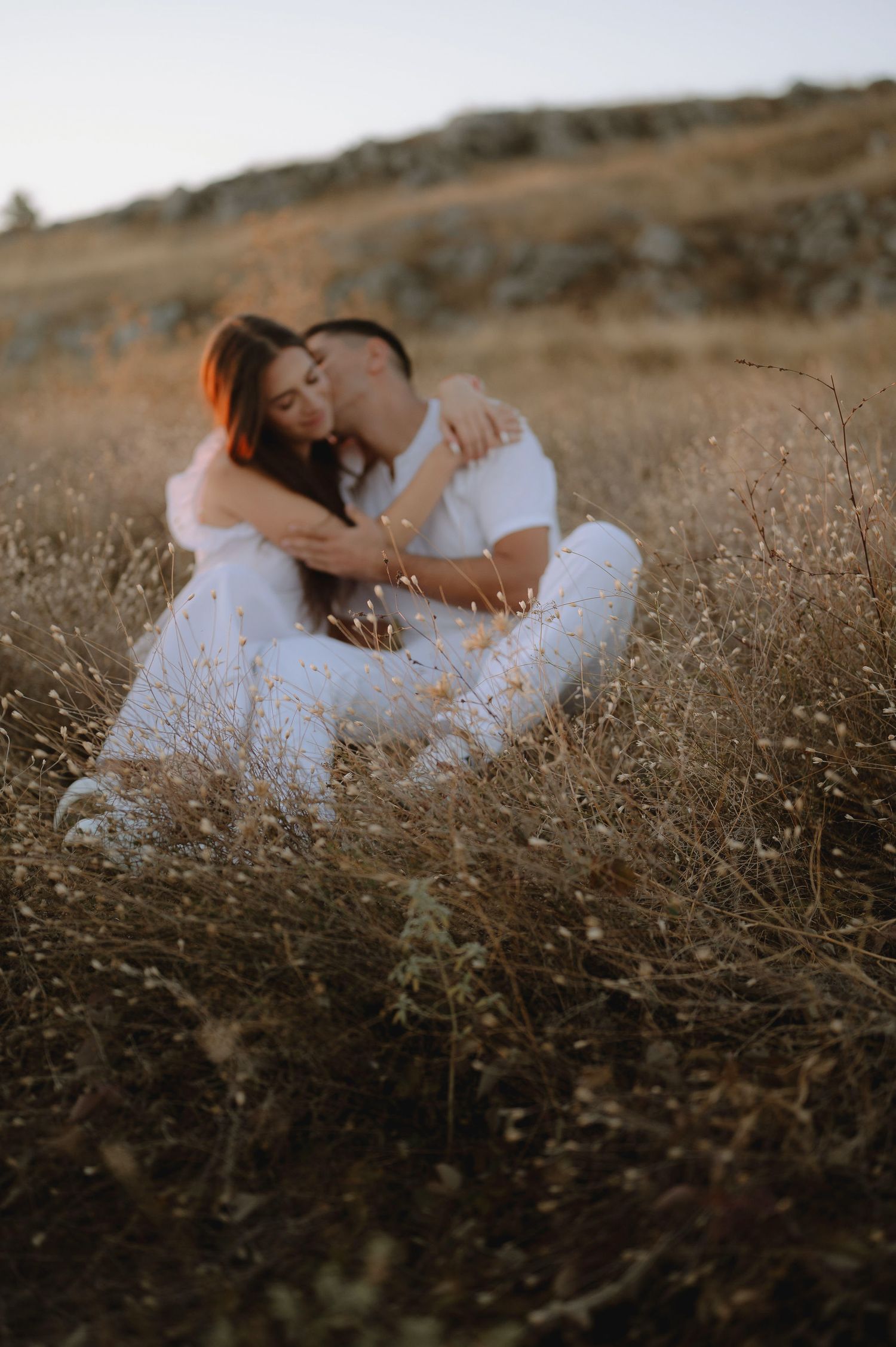 A romantic couple in white clothing embraces while sitting in a golden field during sunset.