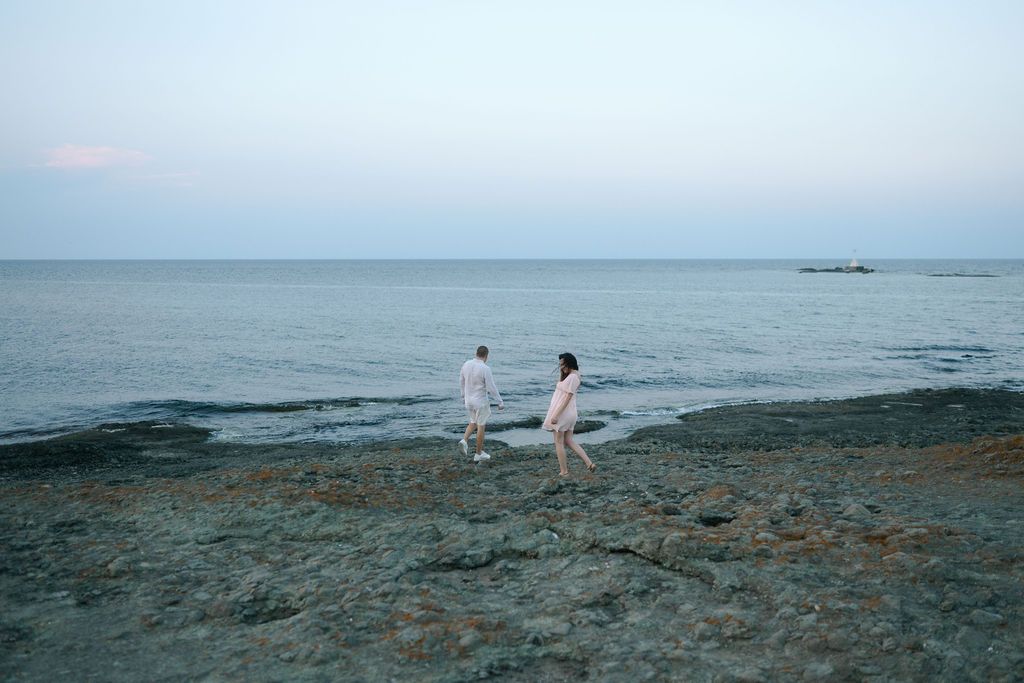 A couple dressed in white strolls on the rocky seaside during sunset with serene waters stretching to the horizon.