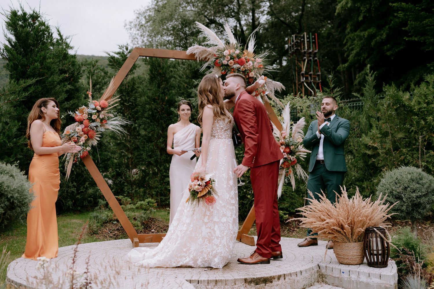 A wedding party poses together in a lush outdoor garden setting with burgundy and orange color accents.