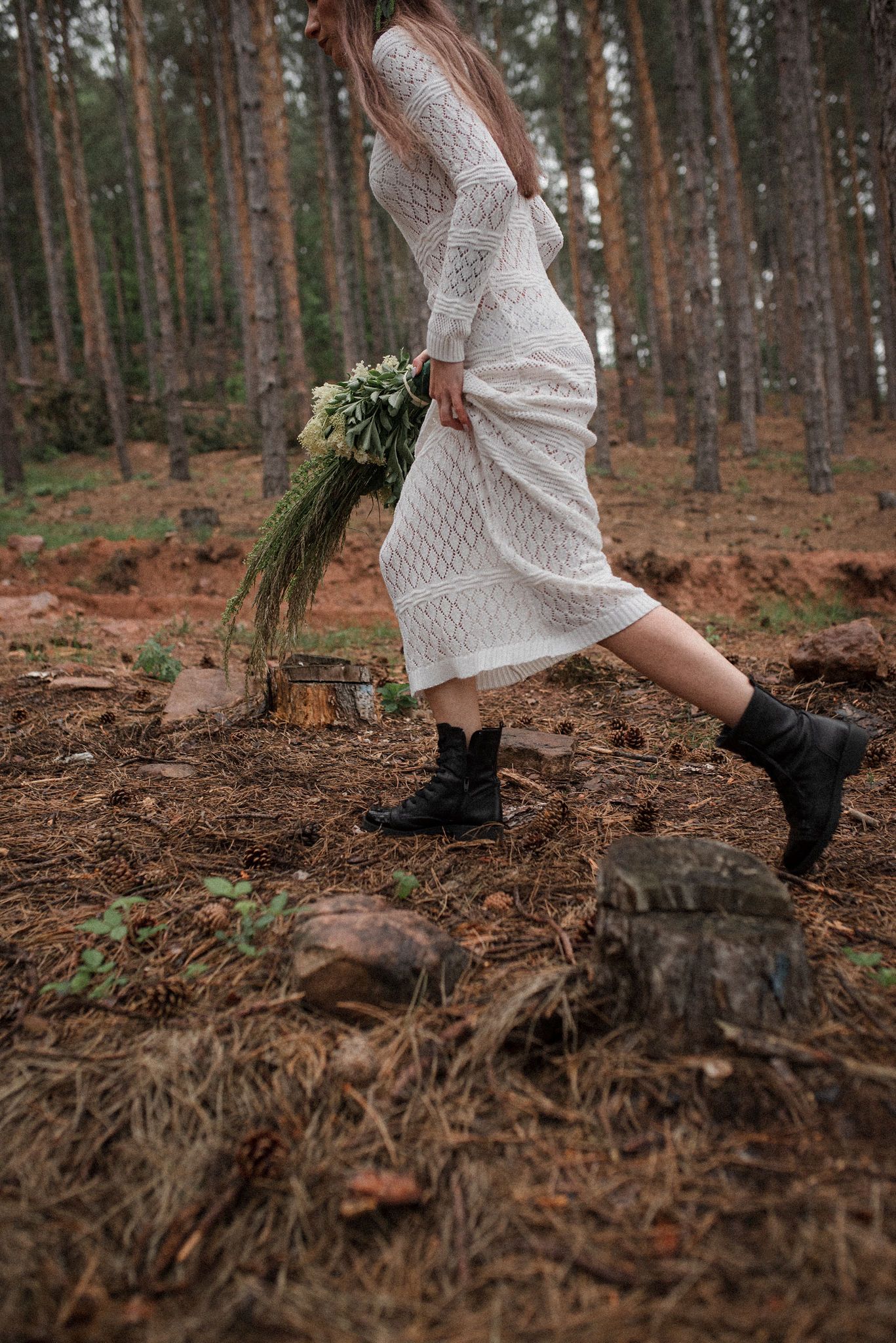A person wearing a white dress and black boots walks through a pine forest with fallen needles on the ground.