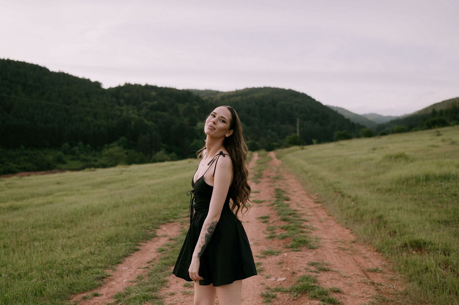 Woman in black dress twirling joyfully on dirt path through green mountain valley.