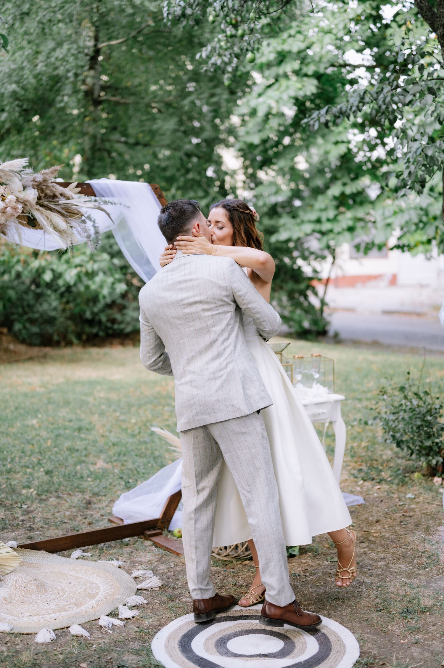 A romantic wedding couple dips and dances together on a circular rug outdoors surrounded by greenery and trees.