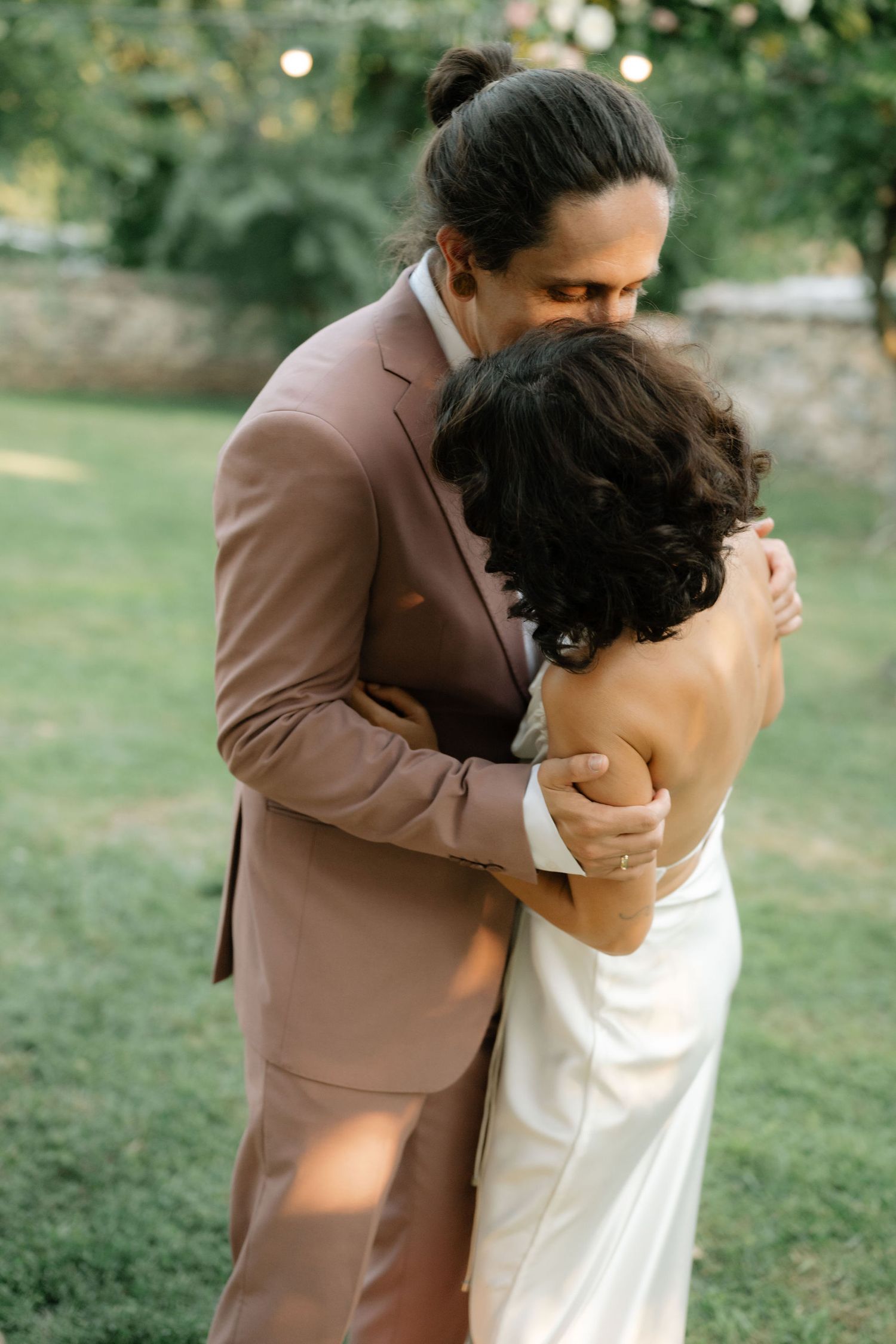 A couple embraces lovingly in an outdoor garden setting wearing brown and white formal attire.