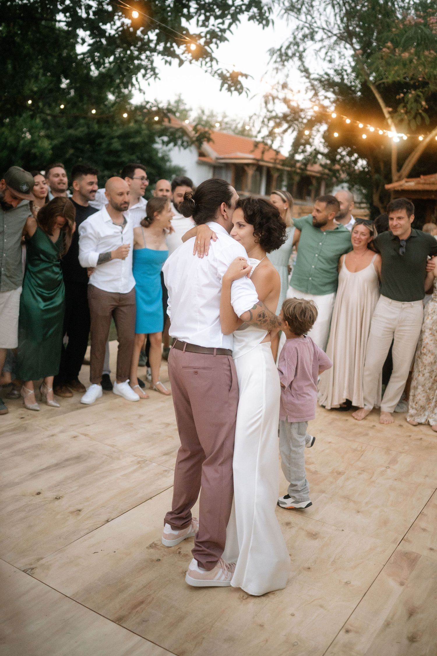Wedding party dances together on an outdoor wooden dance floor during sunset reception with string lights overhead.