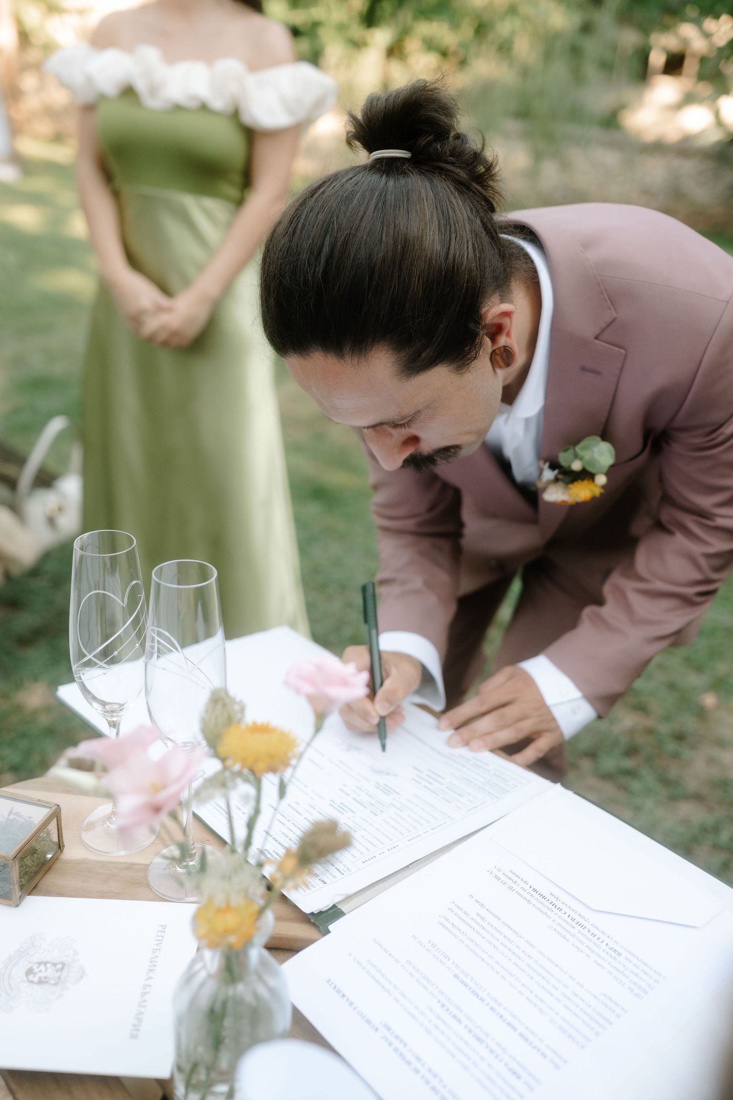 A series of photos showing someone in a brown suit signing wedding documents on a table outdoors with floral decor.