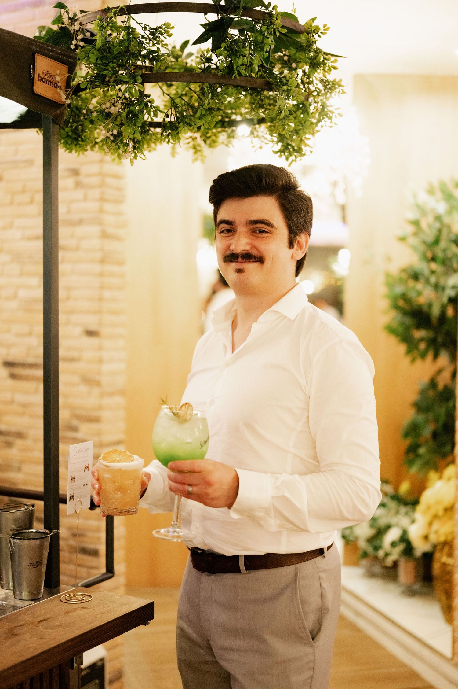 A person in a white dress shirt and dark pants stands in an elegant interior space with greenery and brick walls.