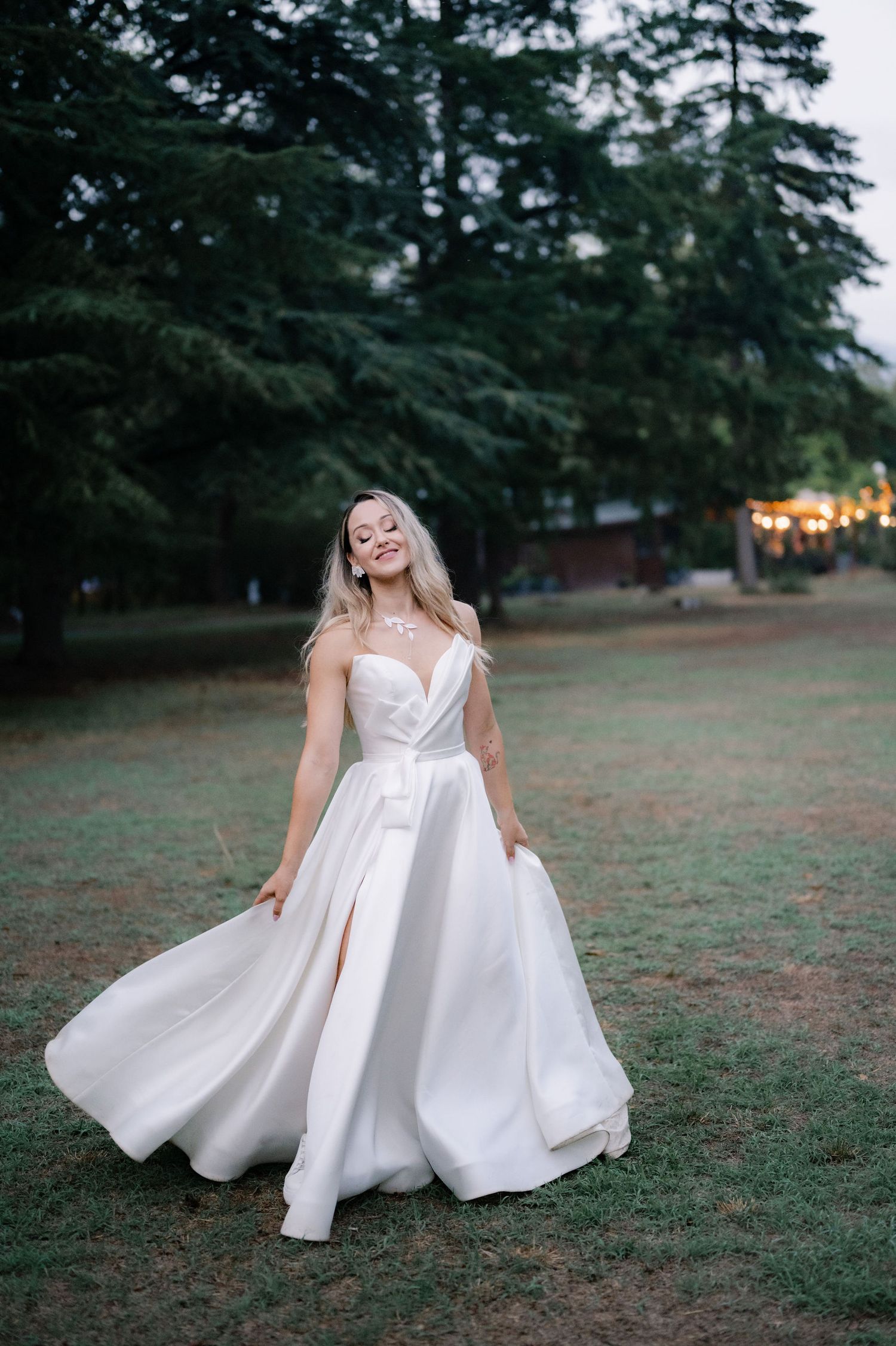 A flowing white wedding dress twirls gracefully in an outdoor setting with trees and string lights in the background.
