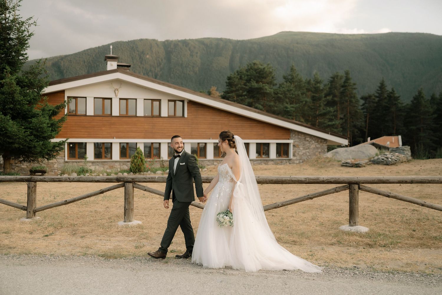 A couple in wedding attire walks together on a path with a mountain lodge and evergreen forest backdrop.