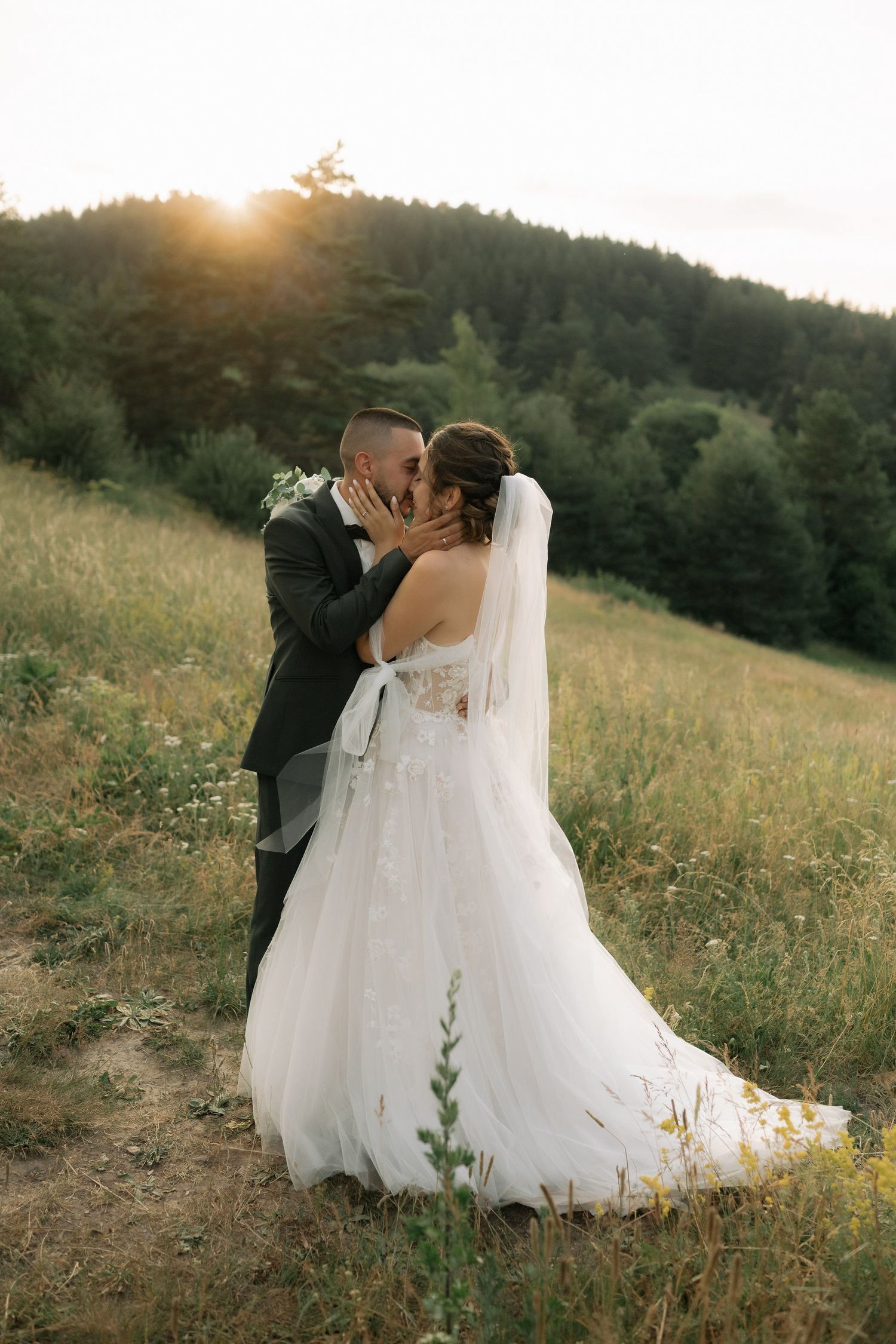 A romantic wedding couple embraces in a golden meadow at sunset with mountains in the background.