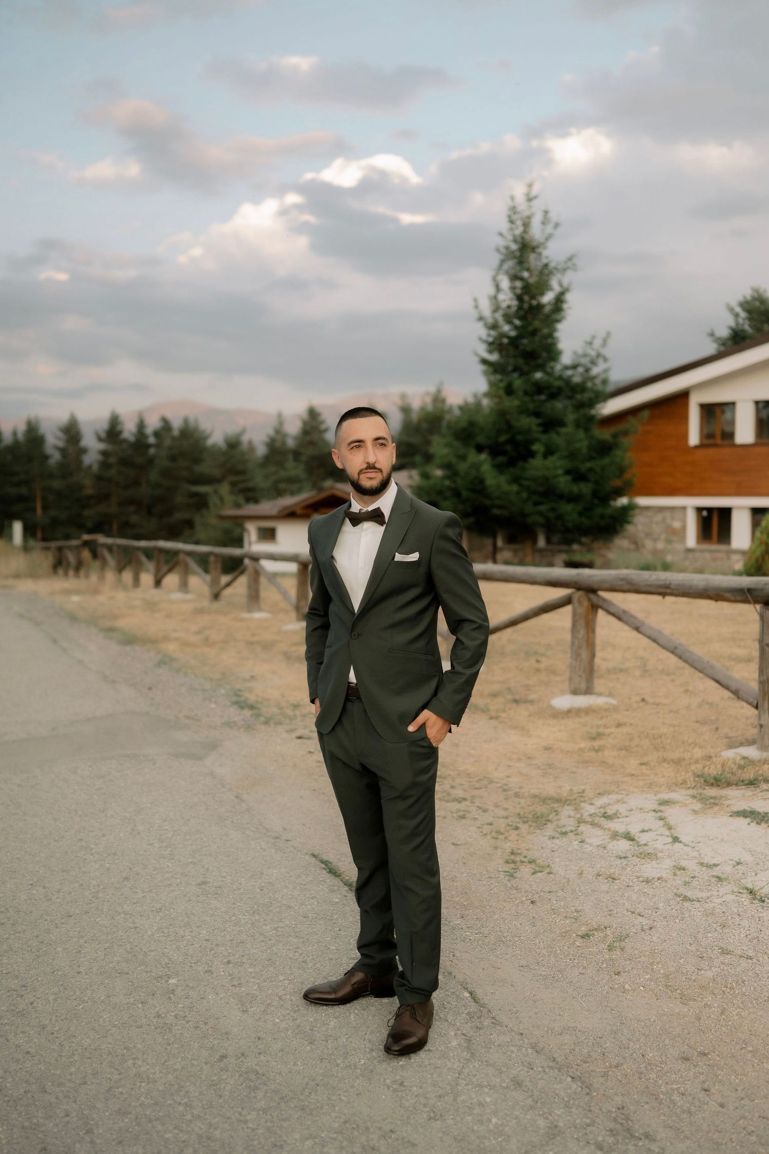 A person in a formal black suit poses on a gravel path with wooden fencing and evergreen trees in the background.