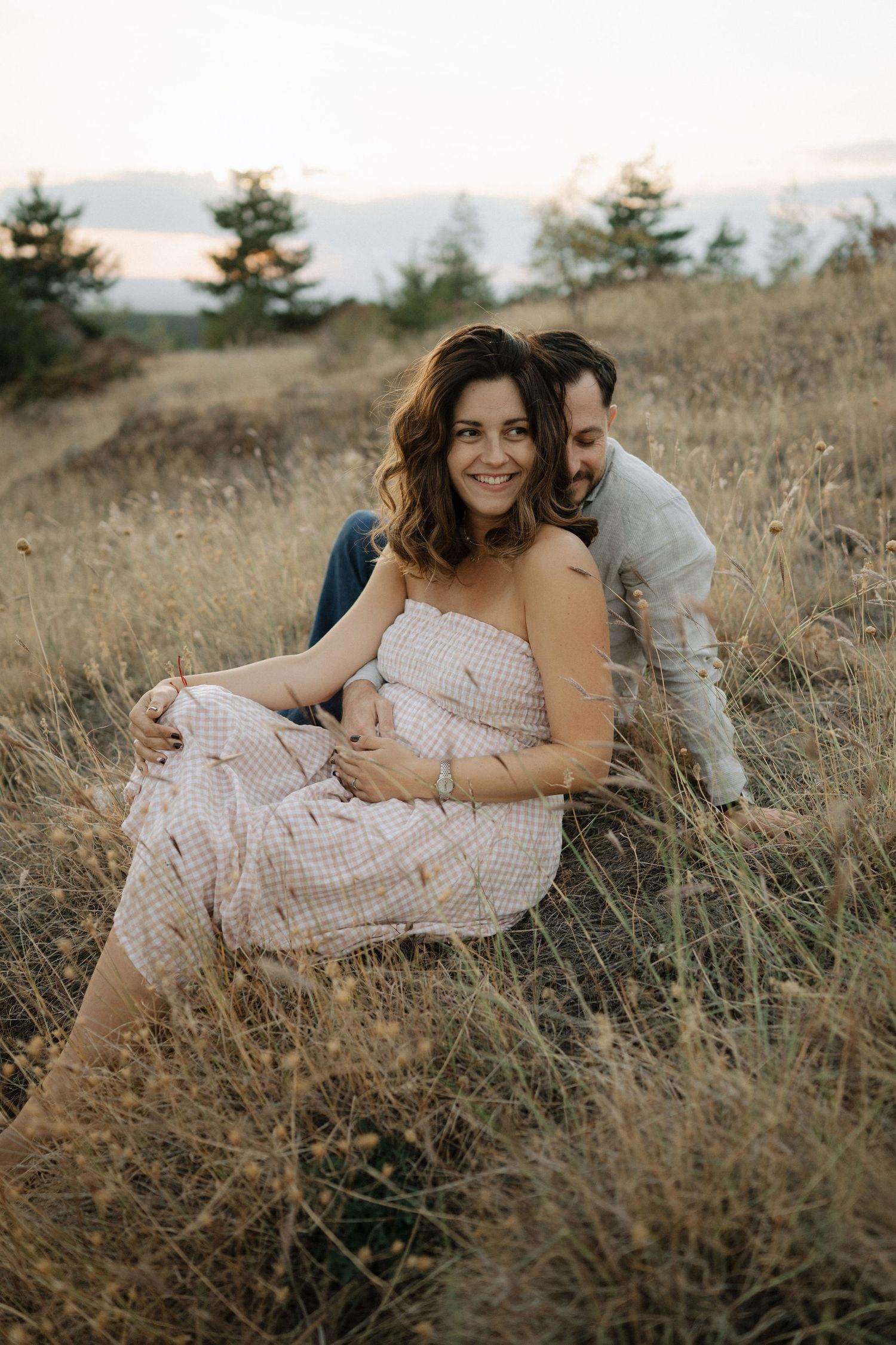 A couple embraces while sitting in a golden field during sunset, creating a romantic outdoor portrait series.