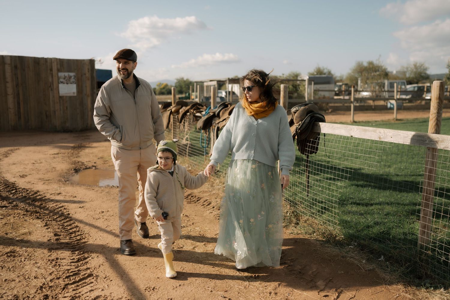 A family walks together along a fence-lined path at a countryside property on a sunny day.