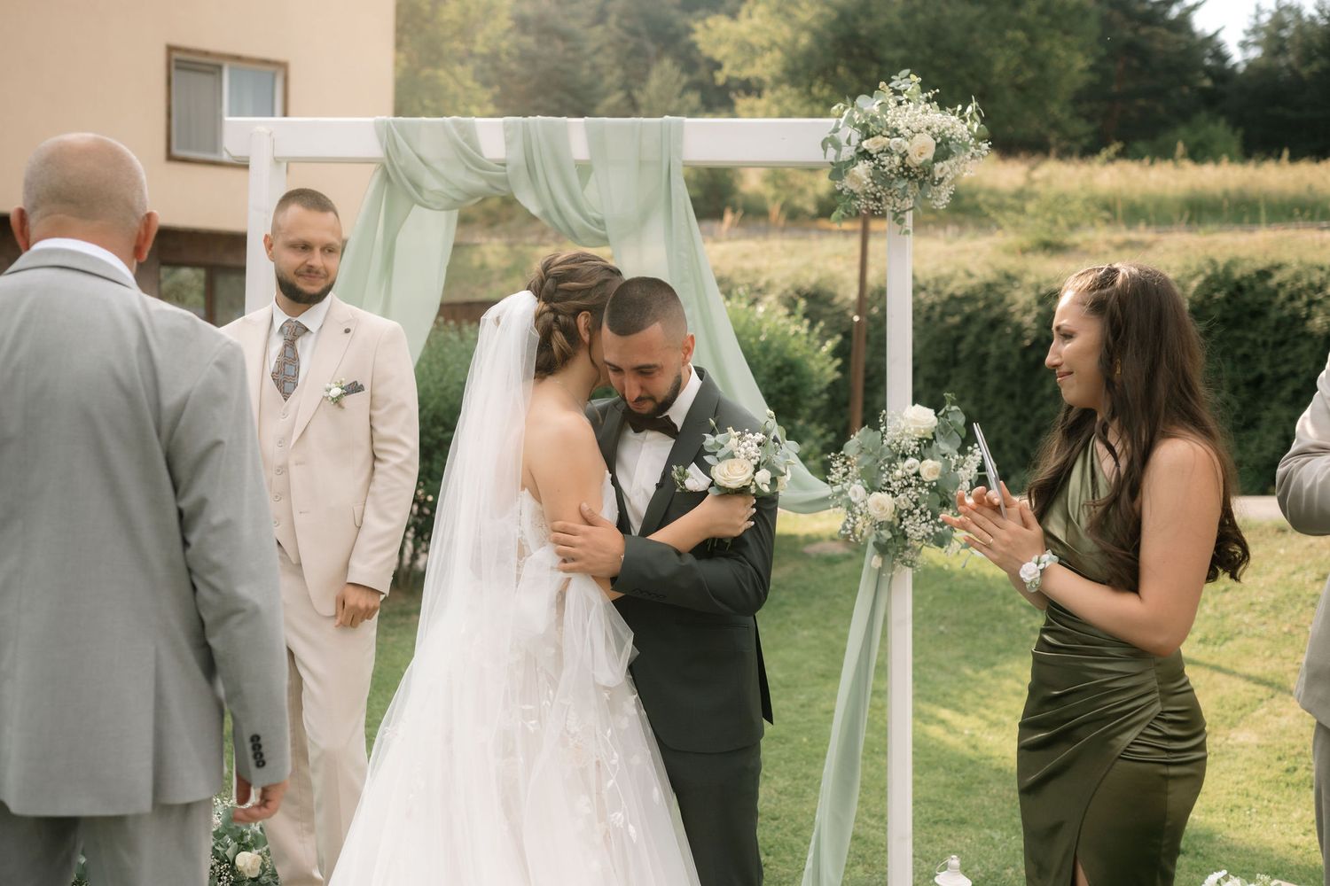 A couple shares an intimate moment during an outdoor wedding ceremony under a floral arch on a sunny day.