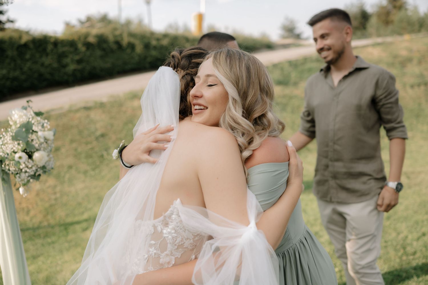A joyful wedding party laughs together in a sunny outdoor setting with natural greenery.