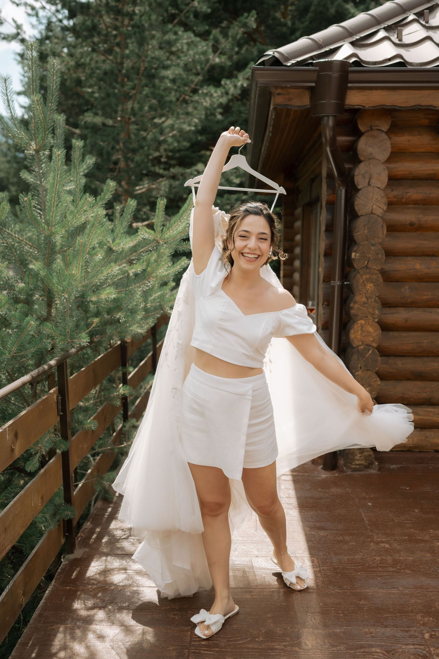 A person in white clothing dances playfully on a rustic wooden cabin porch surrounded by greenery.