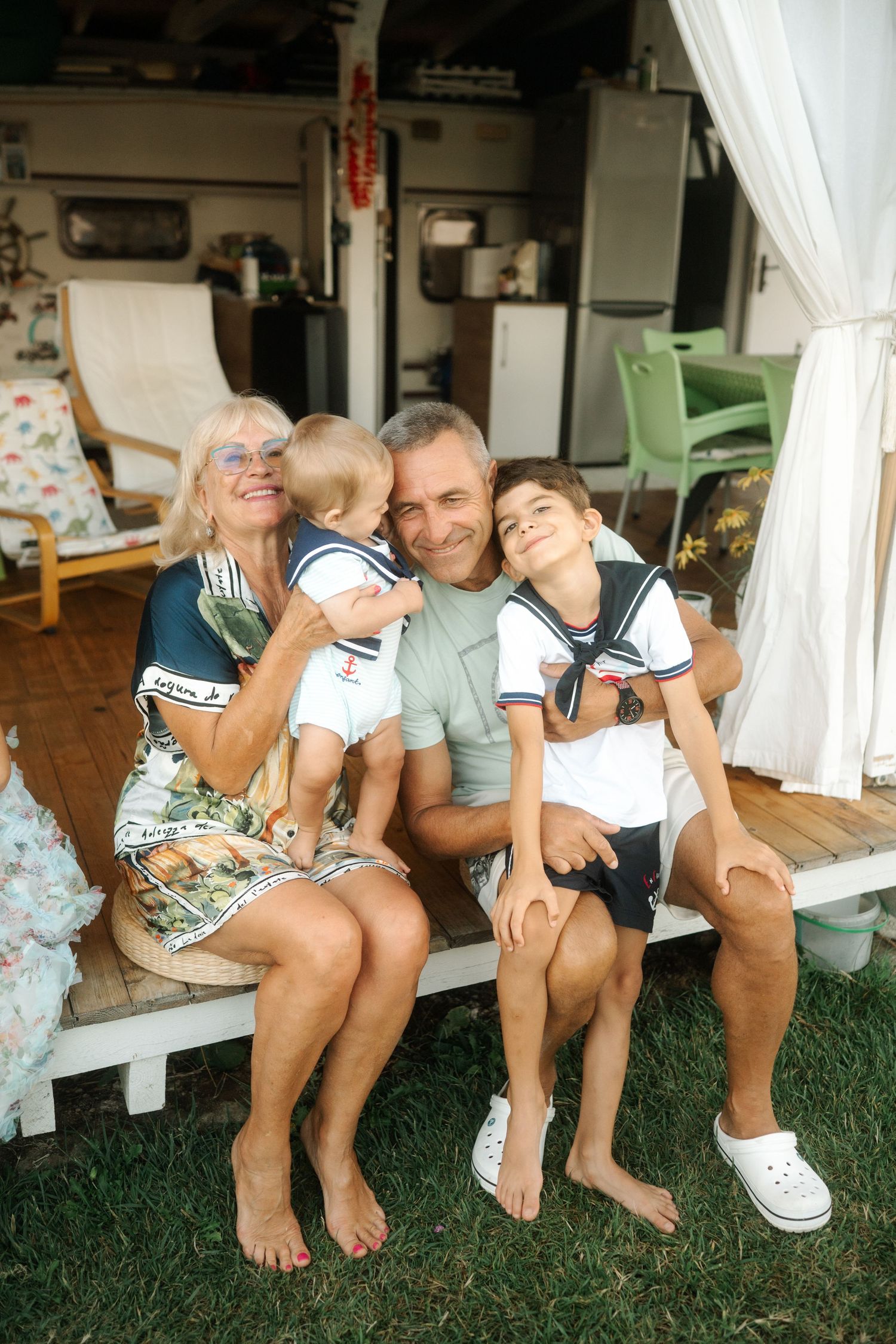 A family sits together on a white porch bench outdoors on a grassy lawn in casual summer attire.