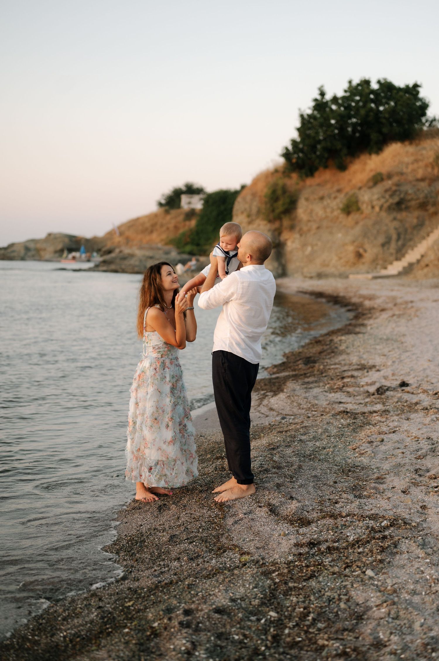A couple dances together on a rocky beach at sunset with water and hills in the background.