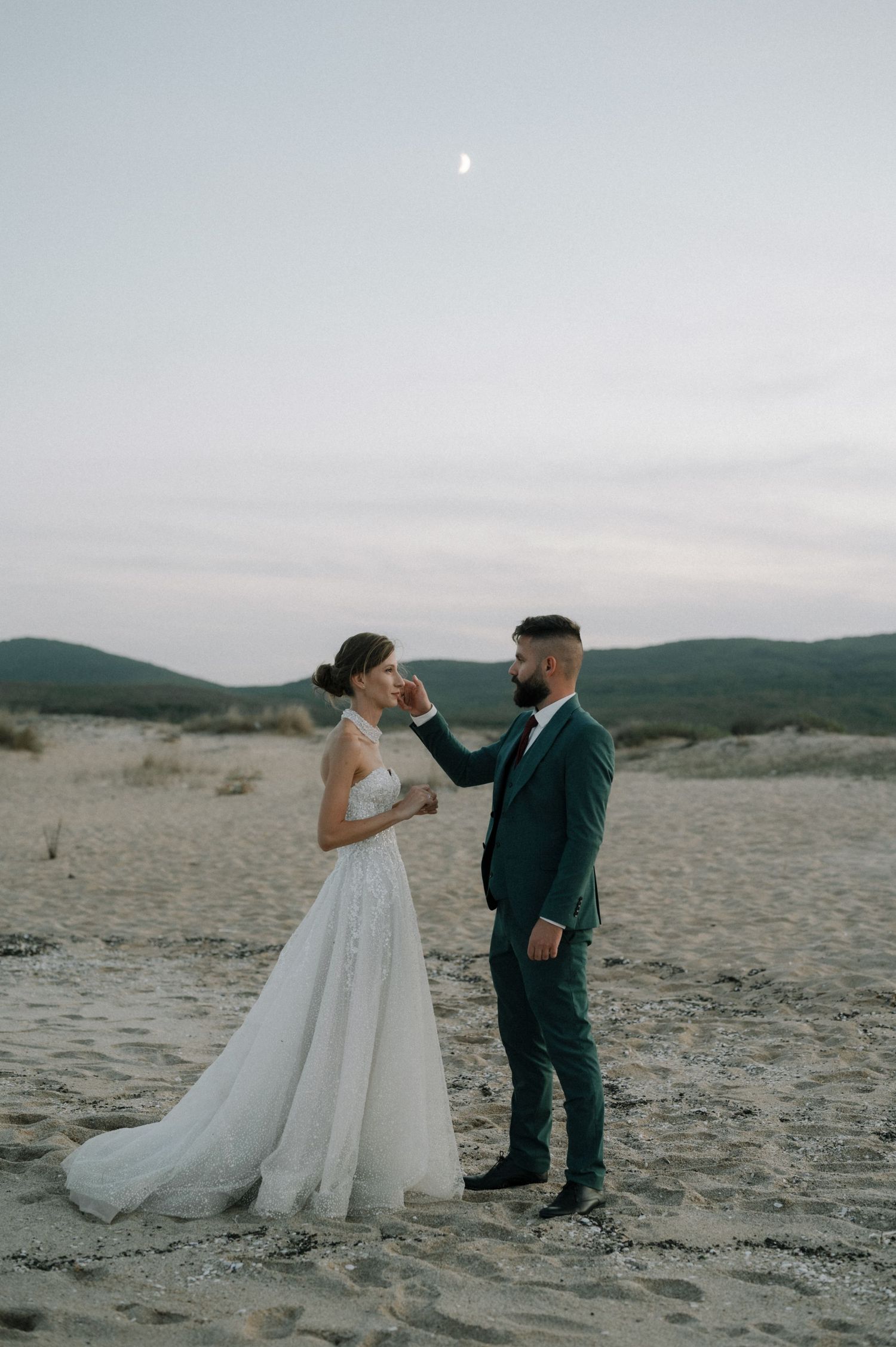 Bride and groom share a romantic moment on sandy beach at dusk with crescent moon and hills in background.